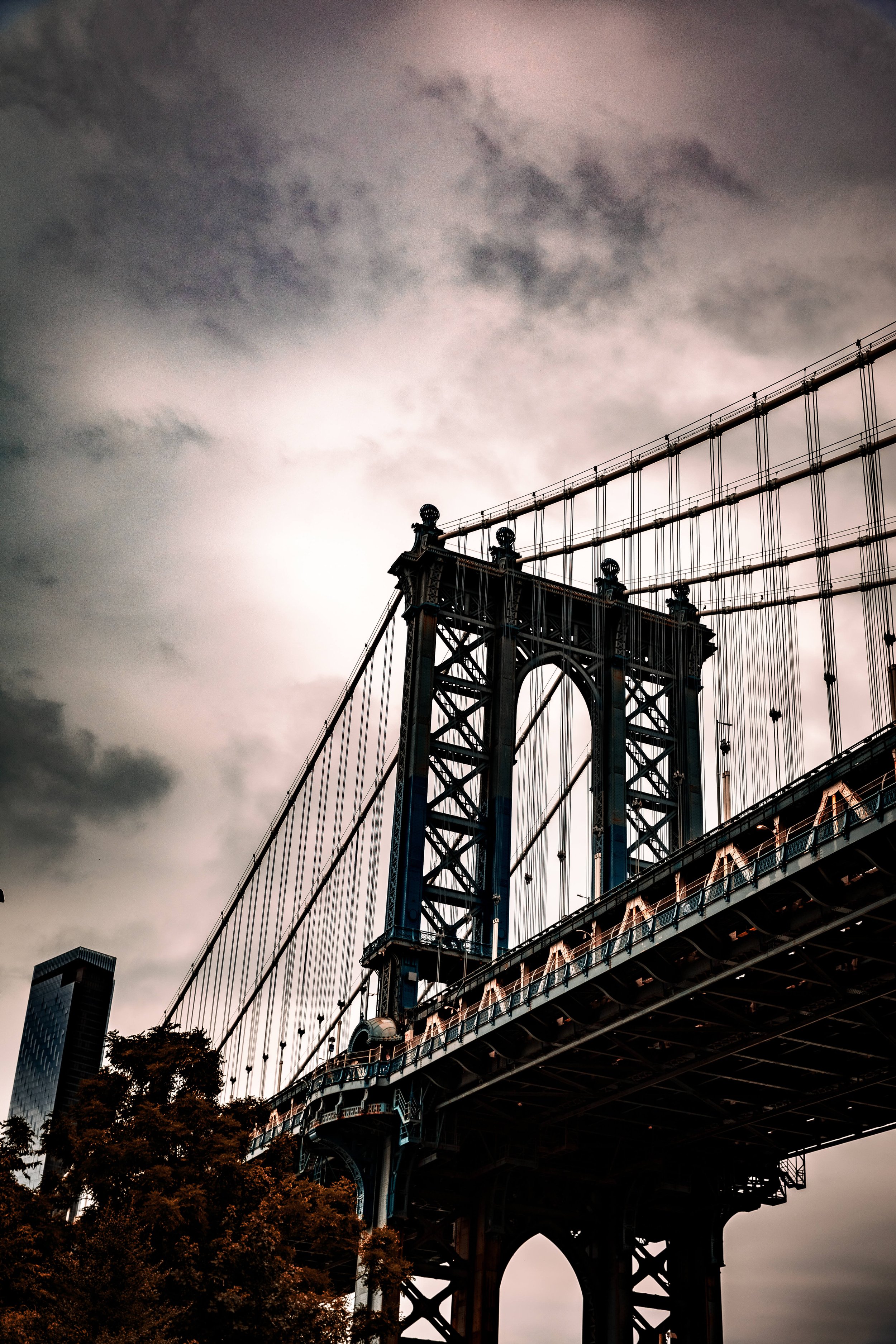 A view of the Brooklyn Bridge against a cloudy sky, with dark metal structural elements and suspension cables, and a faint outline of skyscrapers in the background.