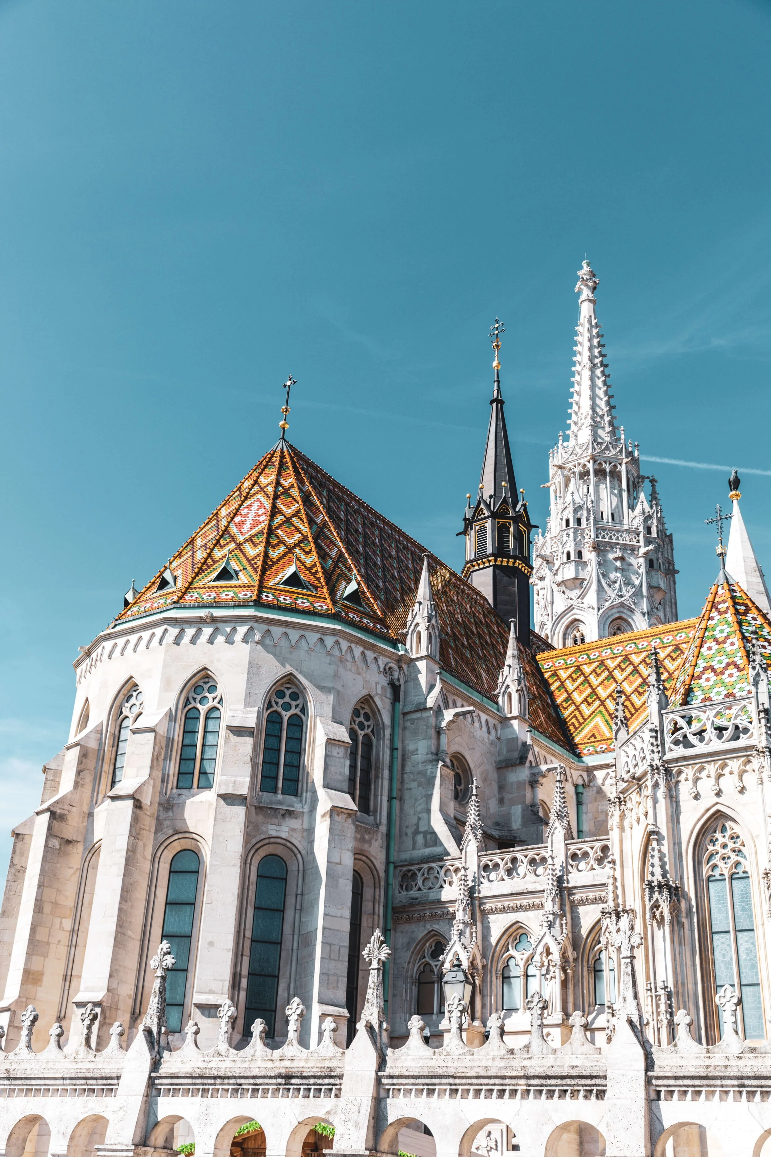 The image shows a close-up view of a gothic-style cathedral with ornate spires, colorful tiled roof, tall arched windows, and intricate stone carvings, set against a bright blue sky.
