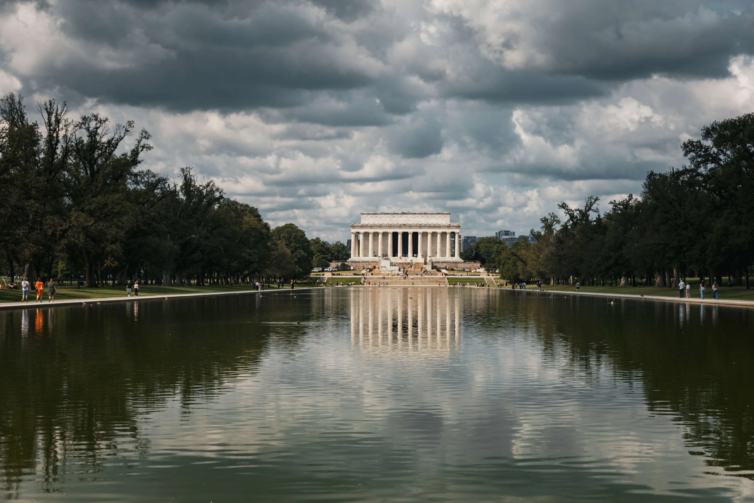 The Lincoln Memorial seen across the Reflecting Pool on a cloudy day, with trees on either side and people walking around.