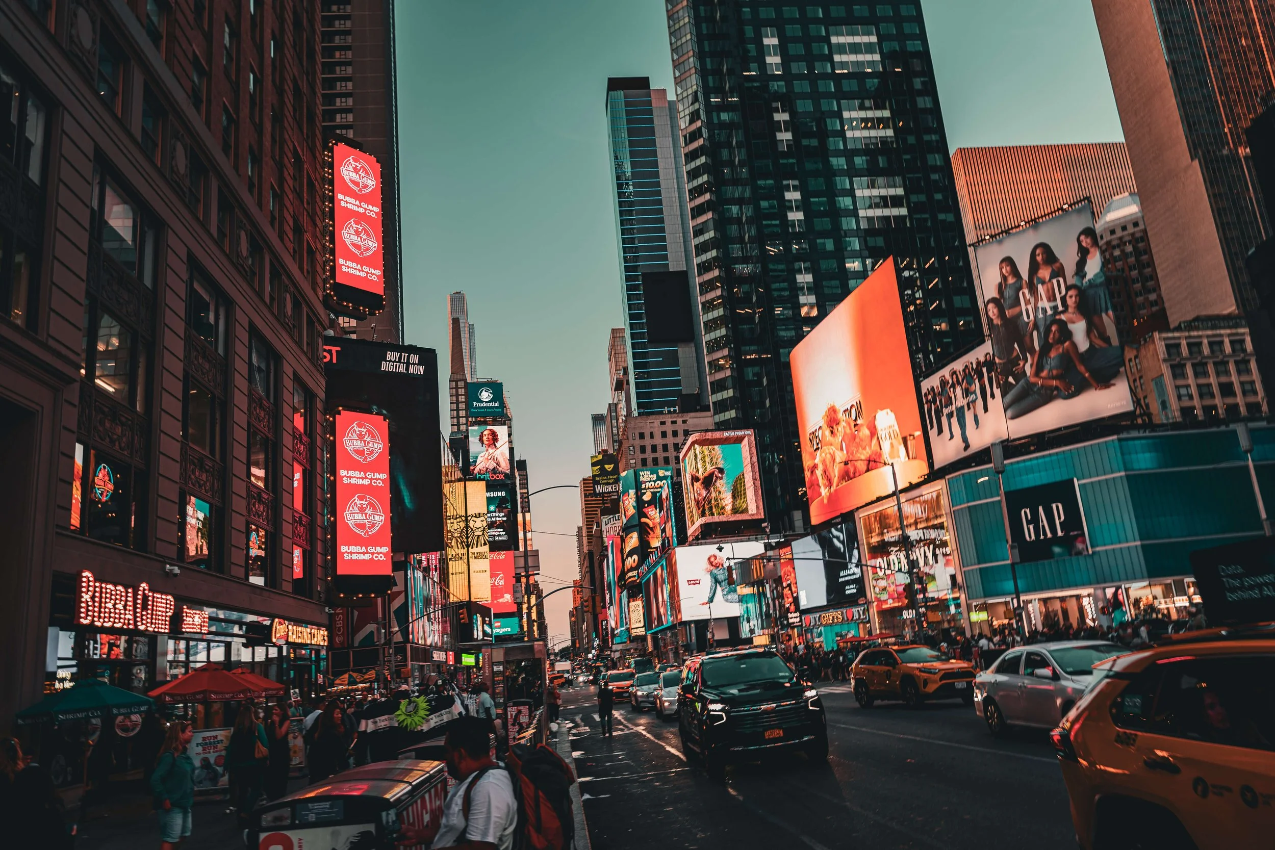 Times Square in New York City at dusk with bright billboards, advertisements, and busy street with cars and pedestrians.