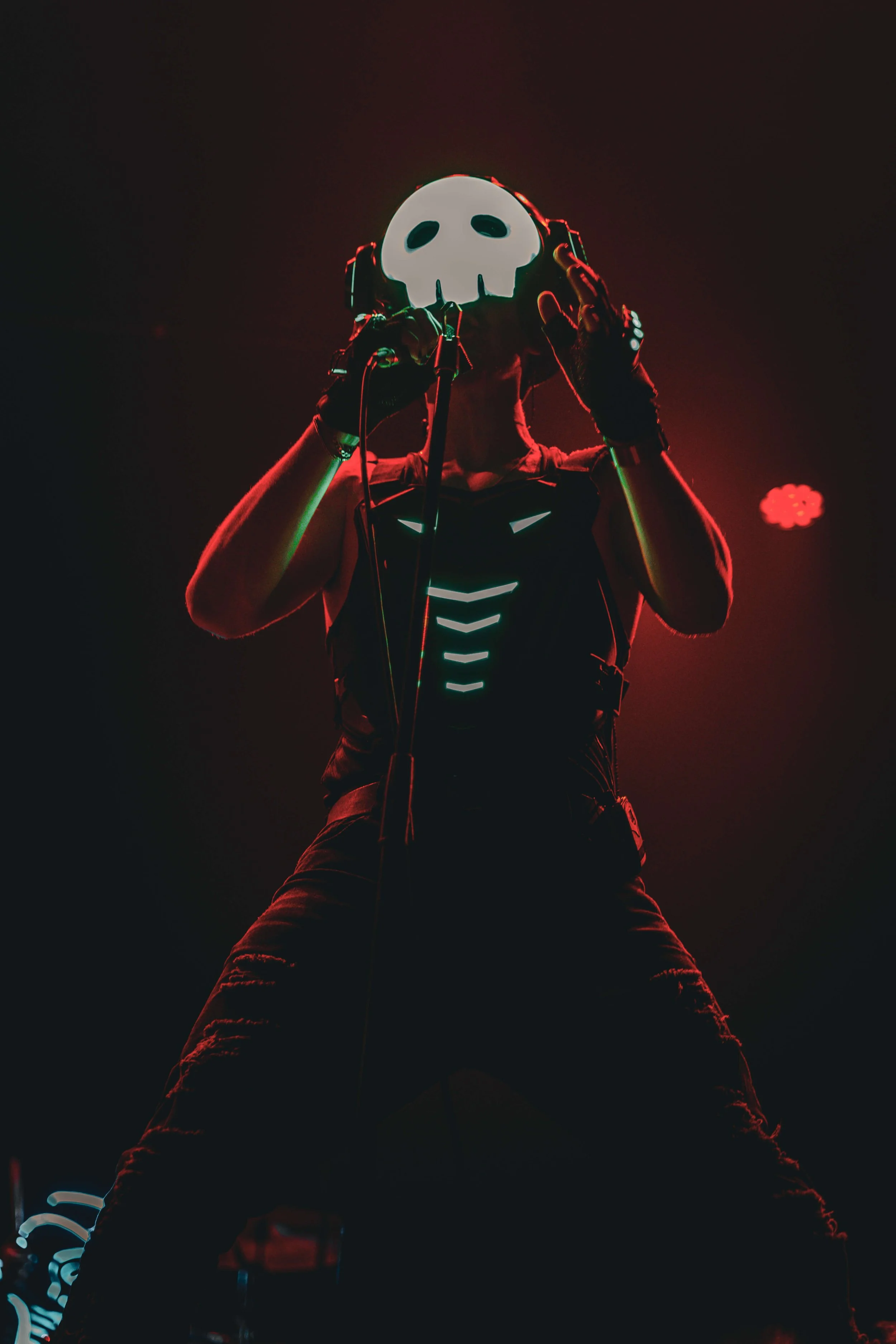 A person wearing a skull mask and black clothing, illuminated by neon red and green lights, in a dark setting, possibly performing or posing.