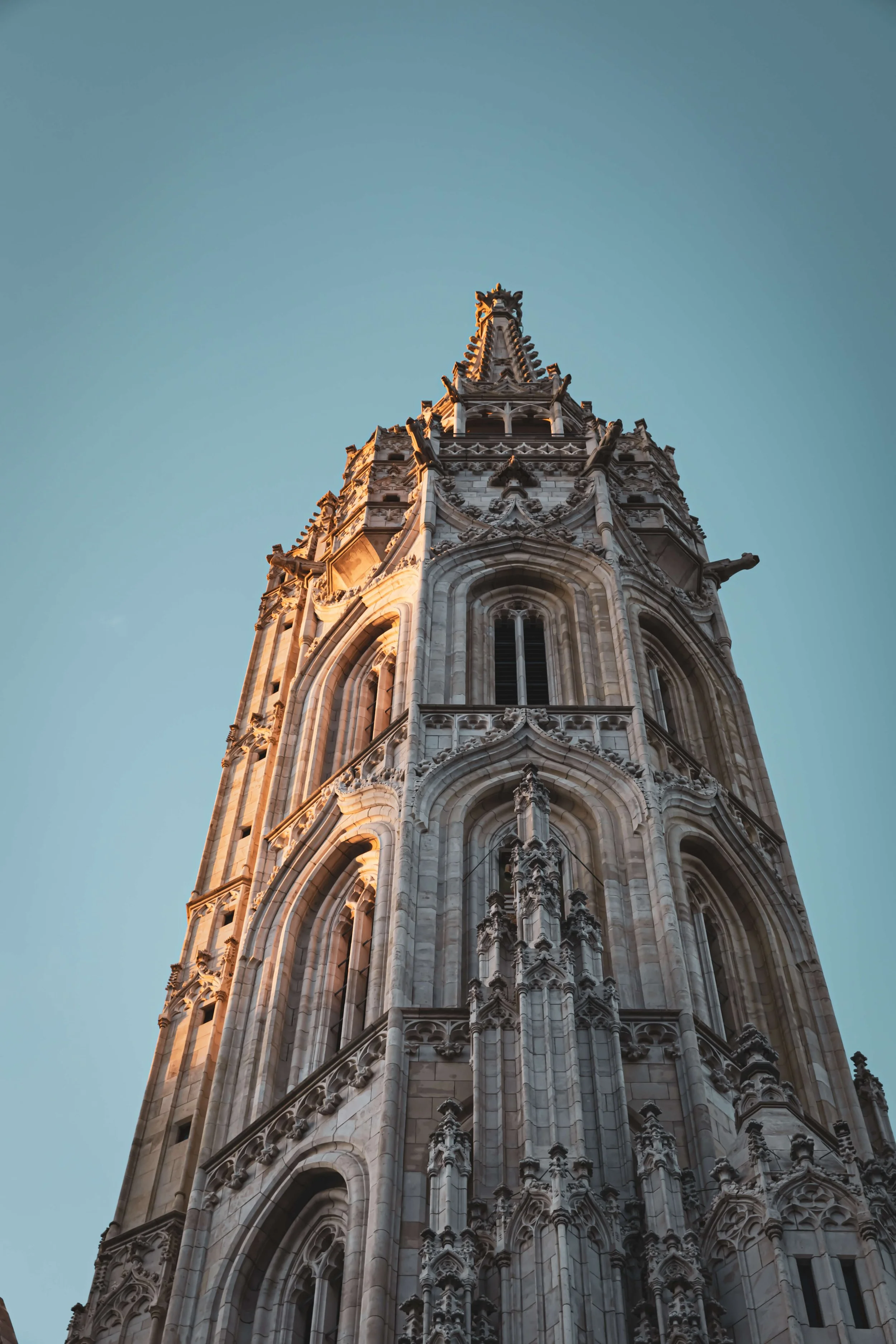 A close-up view of a tall, ornate Gothic-style church tower or spire against a light blue sky, illuminated by warm sunlight.