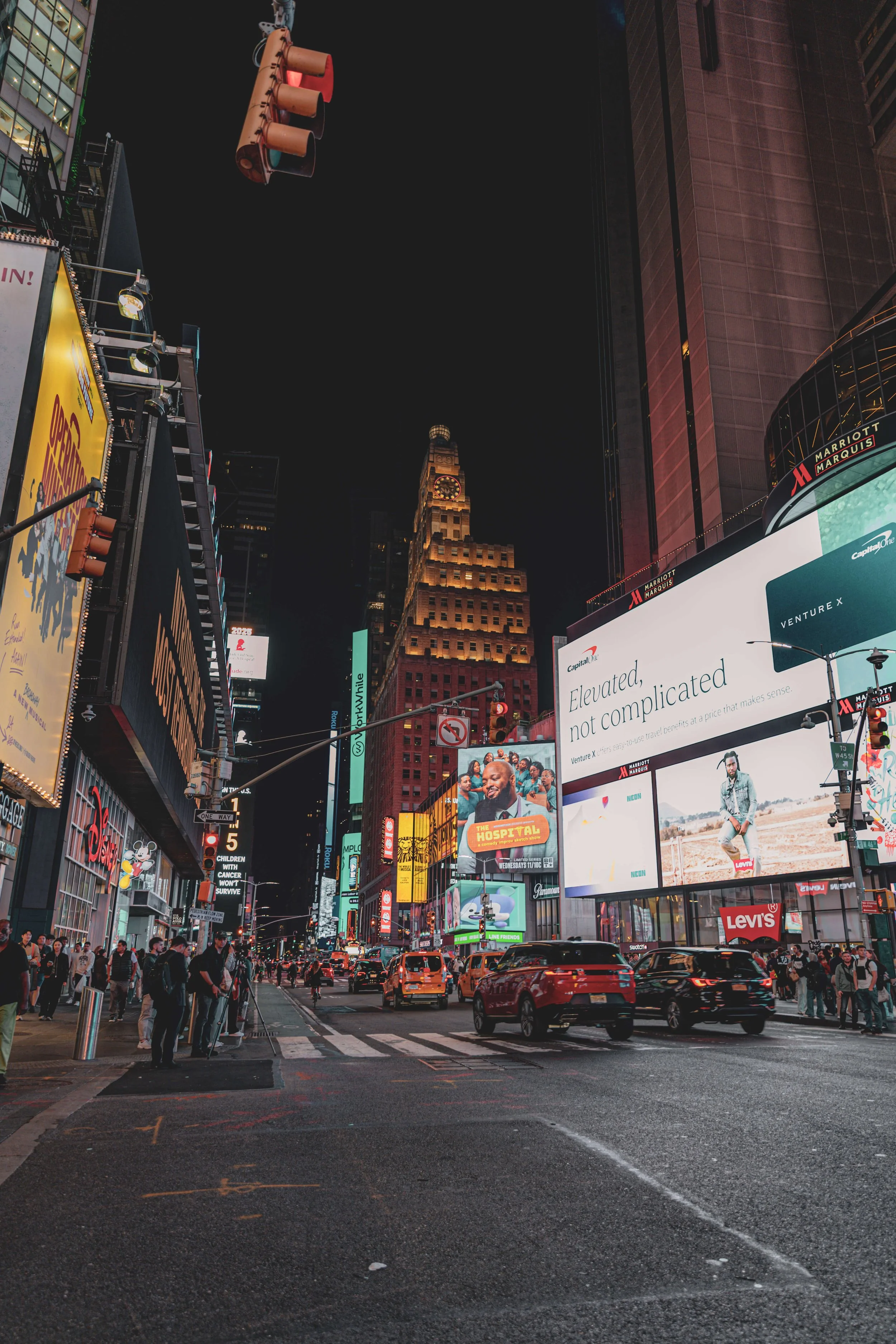 Nighttime in a busy city with illuminated billboards and traffic lights, including the iconic clock tower on a tall building and several cars and pedestrians.