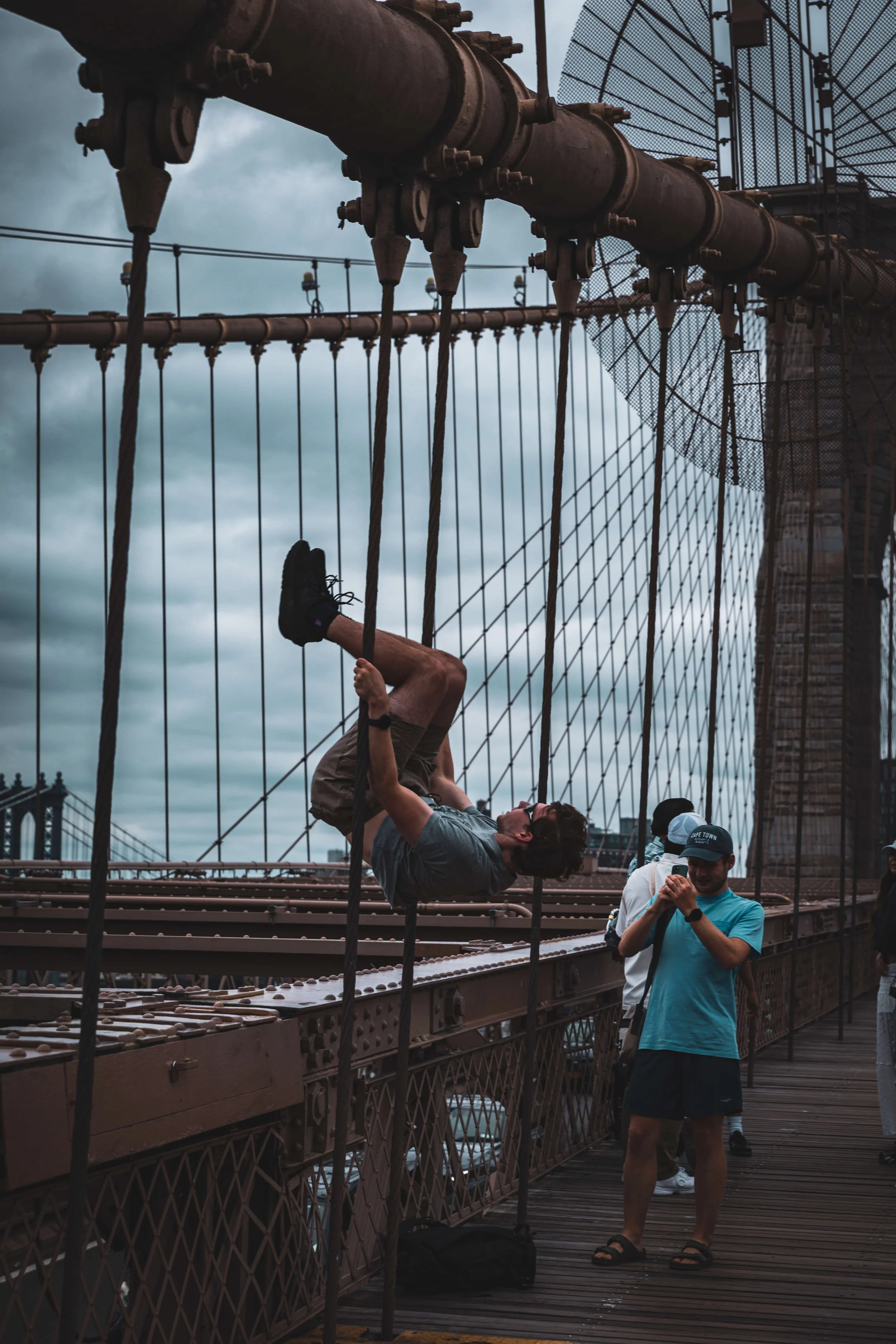 Man hanging upside down on the Brooklyn Bridge with a cloudy sky in the background, while others stand nearby taking photos.