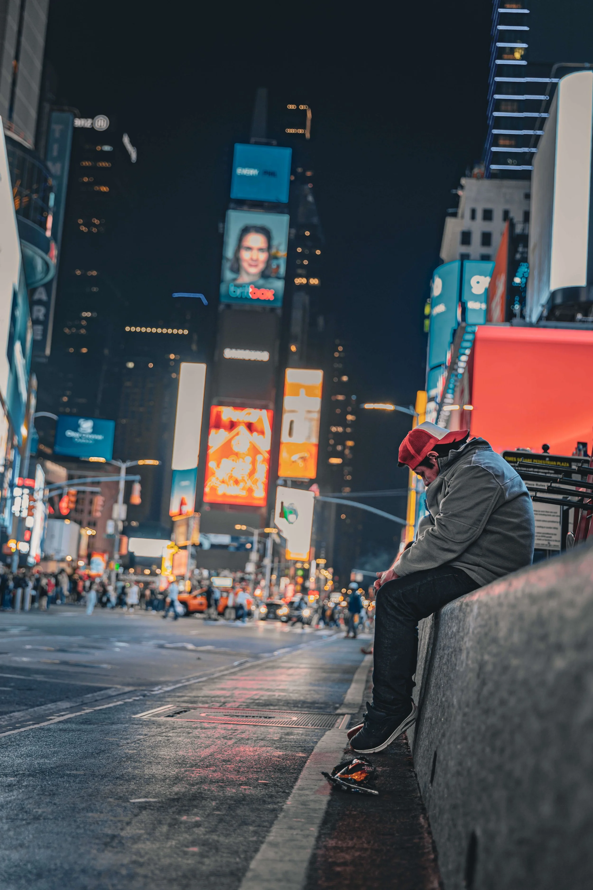 A young man sitting on a concrete ledge at night in Times Square, New York City, with digital billboards and bright city lights in the background.