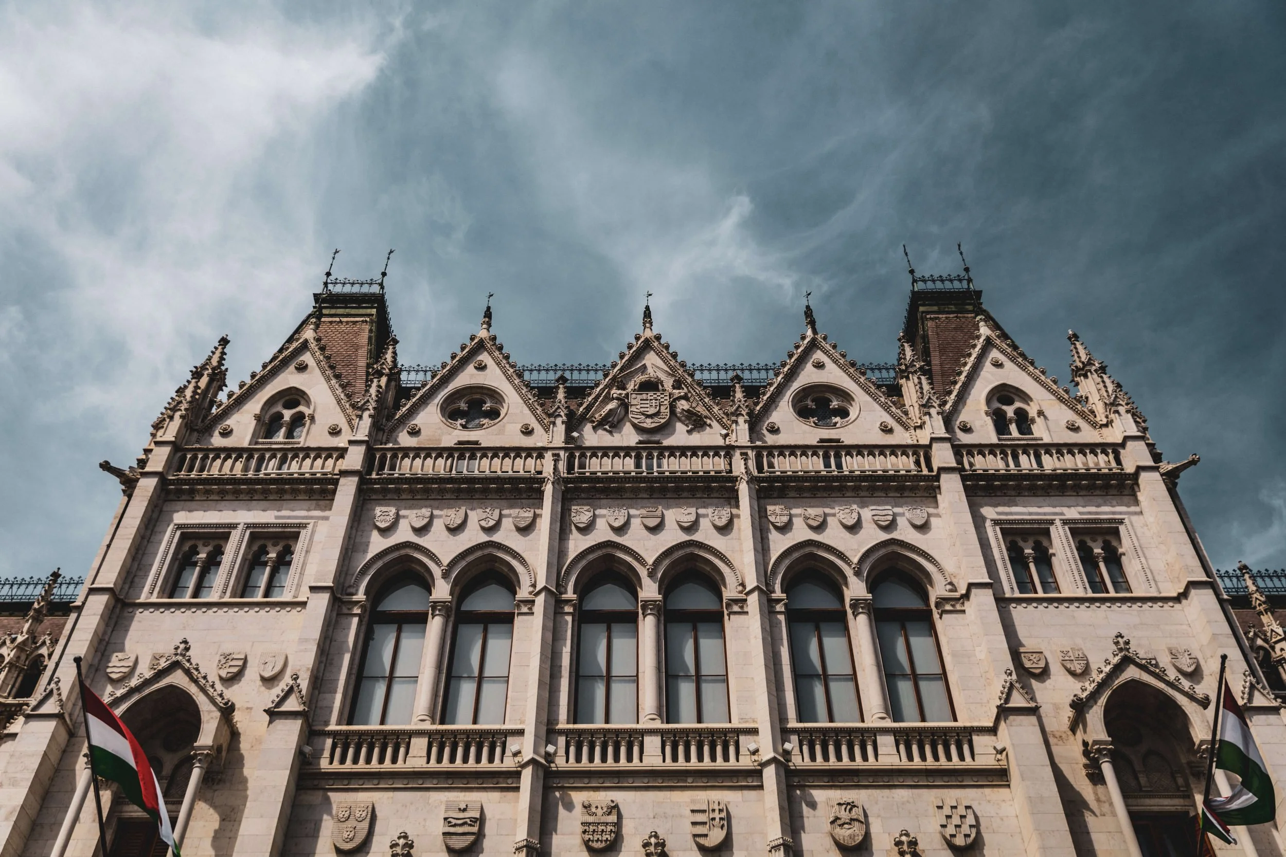 A historic Gothic-style building with decorative elements and multiple coat of arms shields, flags, and ornate spires on the roof against a cloudy sky.