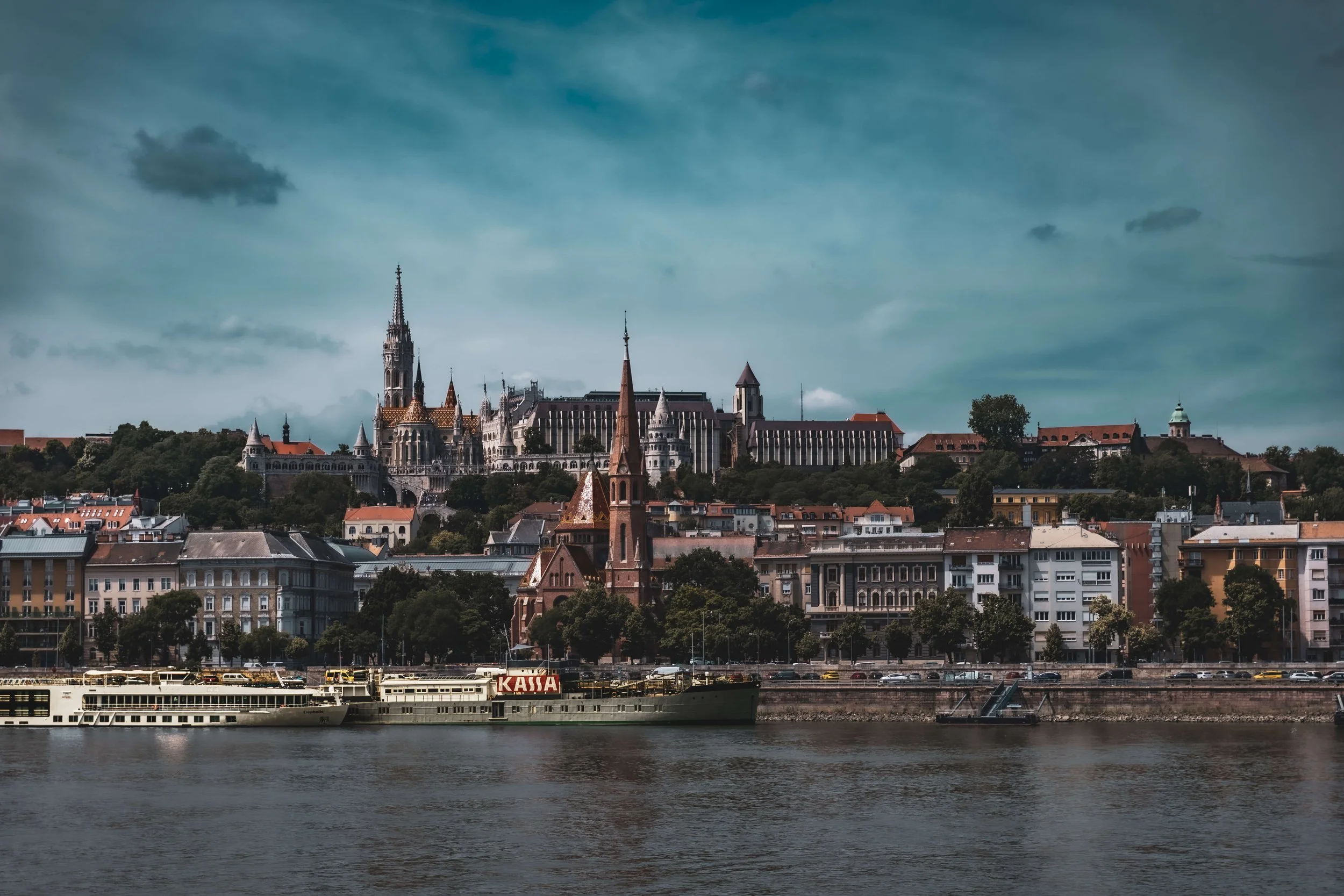 View of a city skyline with historic buildings, a river in the foreground, and a boat docked at the riverbank. The sky is partly cloudy with a few small clouds.