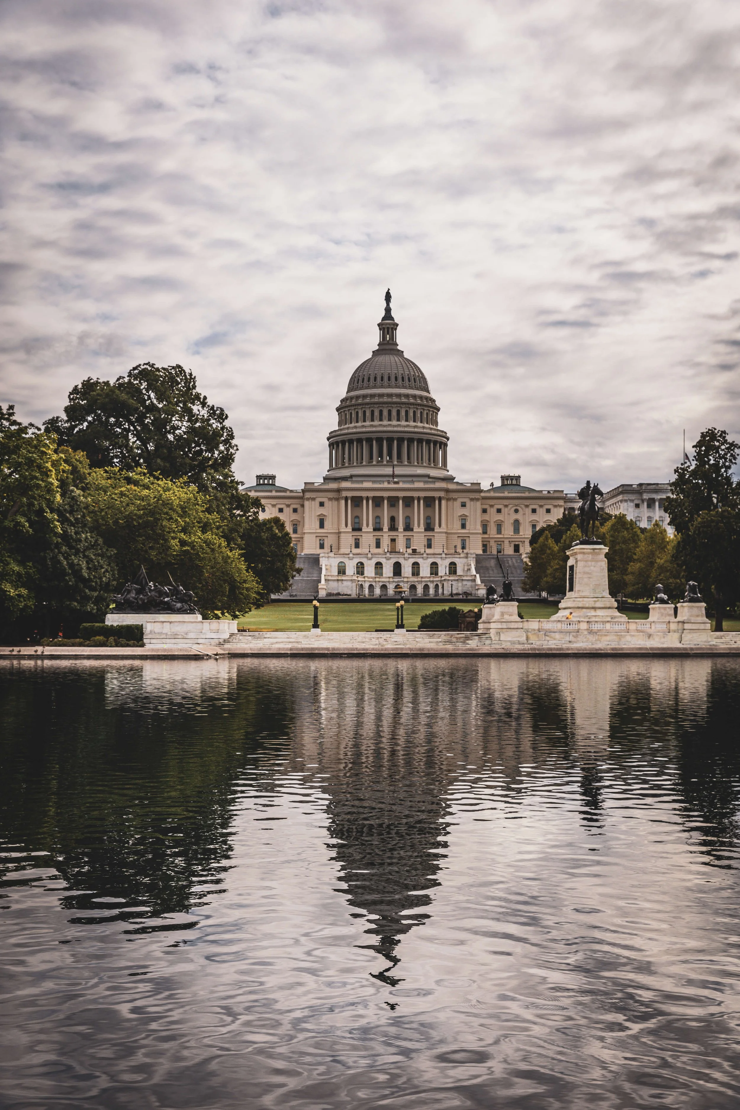 The United States Capitol building in Washington D.C., viewed from the reflecting pond with trees on either side and cloudy sky overhead.
