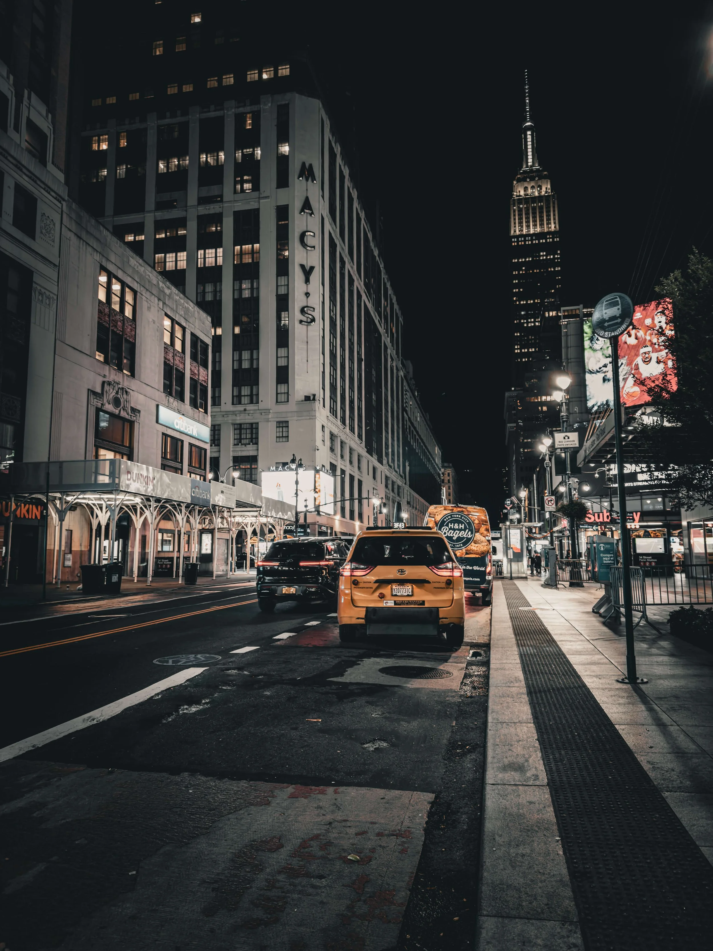 Nighttime city street scene with parked cars, illuminated buildings, and the Empire State Building in the background.