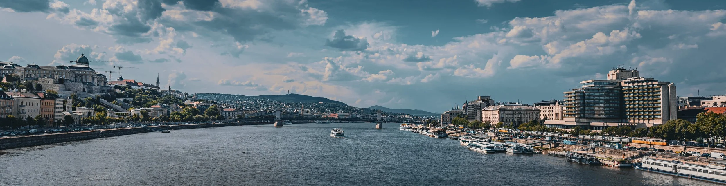 Cityscape of a river with boats, historic and modern buildings along the water, and a cloudy sky.