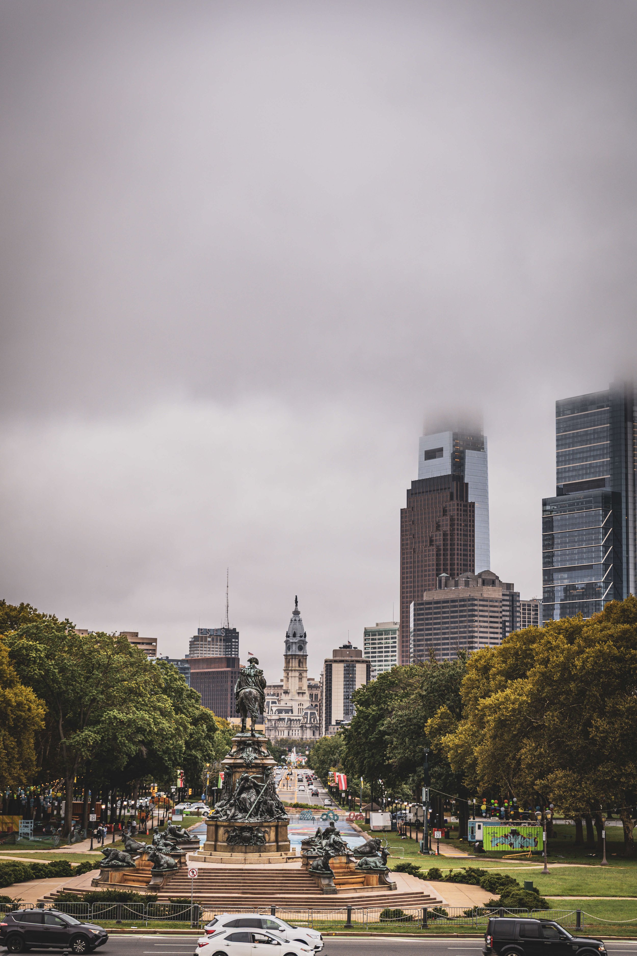 A city park with a large fountain in the foreground, surrounded by trees with yellow-green leaves, and city skyscrapers in the background, some with clouds touching their tops.