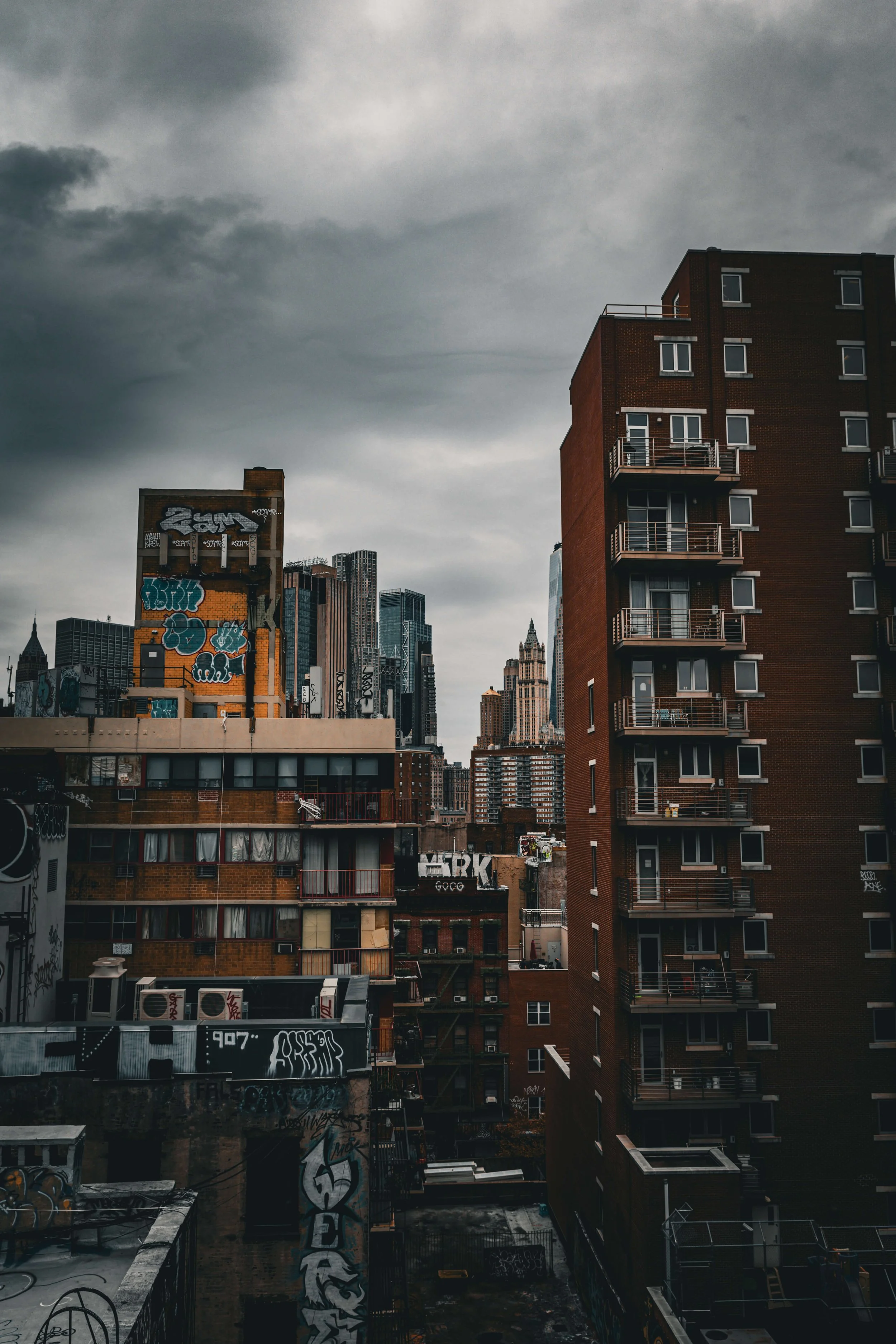 Overcast cityscape featuring tall buildings, some with graffiti on brick exteriors, in a dense urban area under dark, cloudy skies.