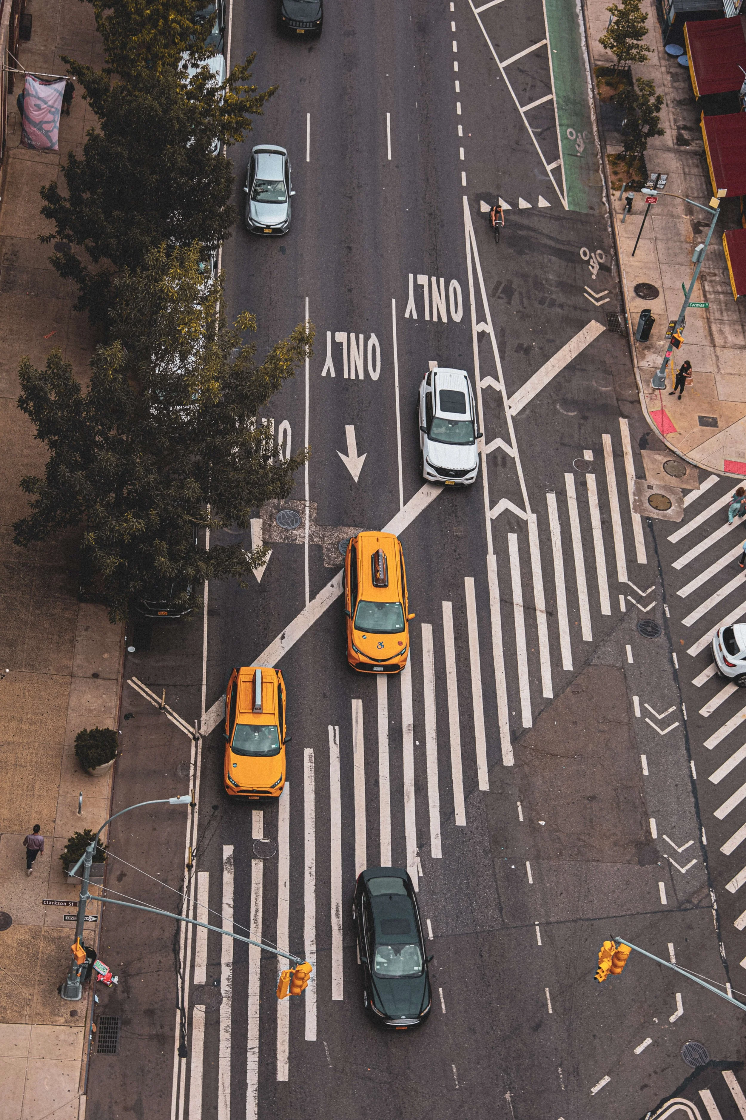 Aerial view of a city intersection showing streets with parking lanes, crosswalks, traffic signals, and cars including two yellow taxis, a black car, and a white car. Pedestrians are on the sidewalks, and a tree is partially blocking the view.