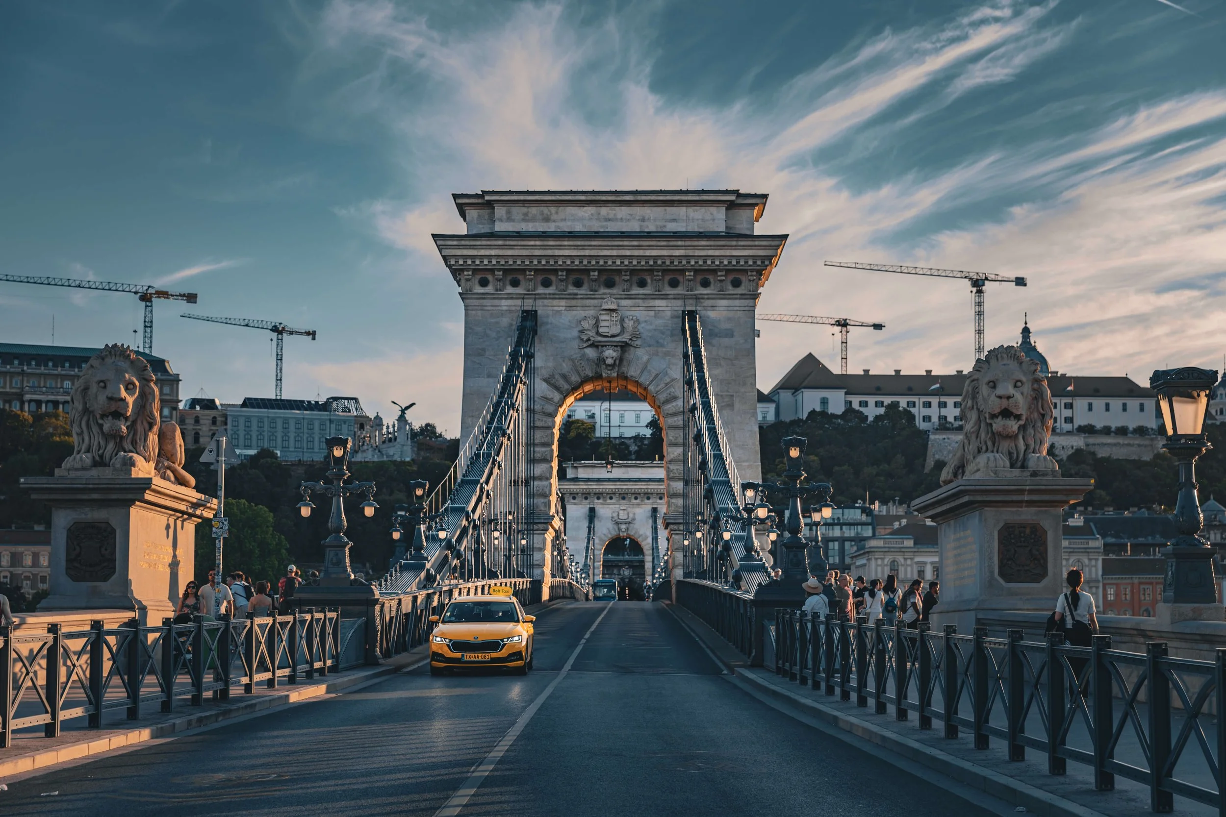 Looking across a bridge with lion statues at each end towards a historic stone archway. The bridge is bustling with pedestrians and a yellow taxi car is driving on it. Construction cranes and buildings are visible in the background under a partly clo