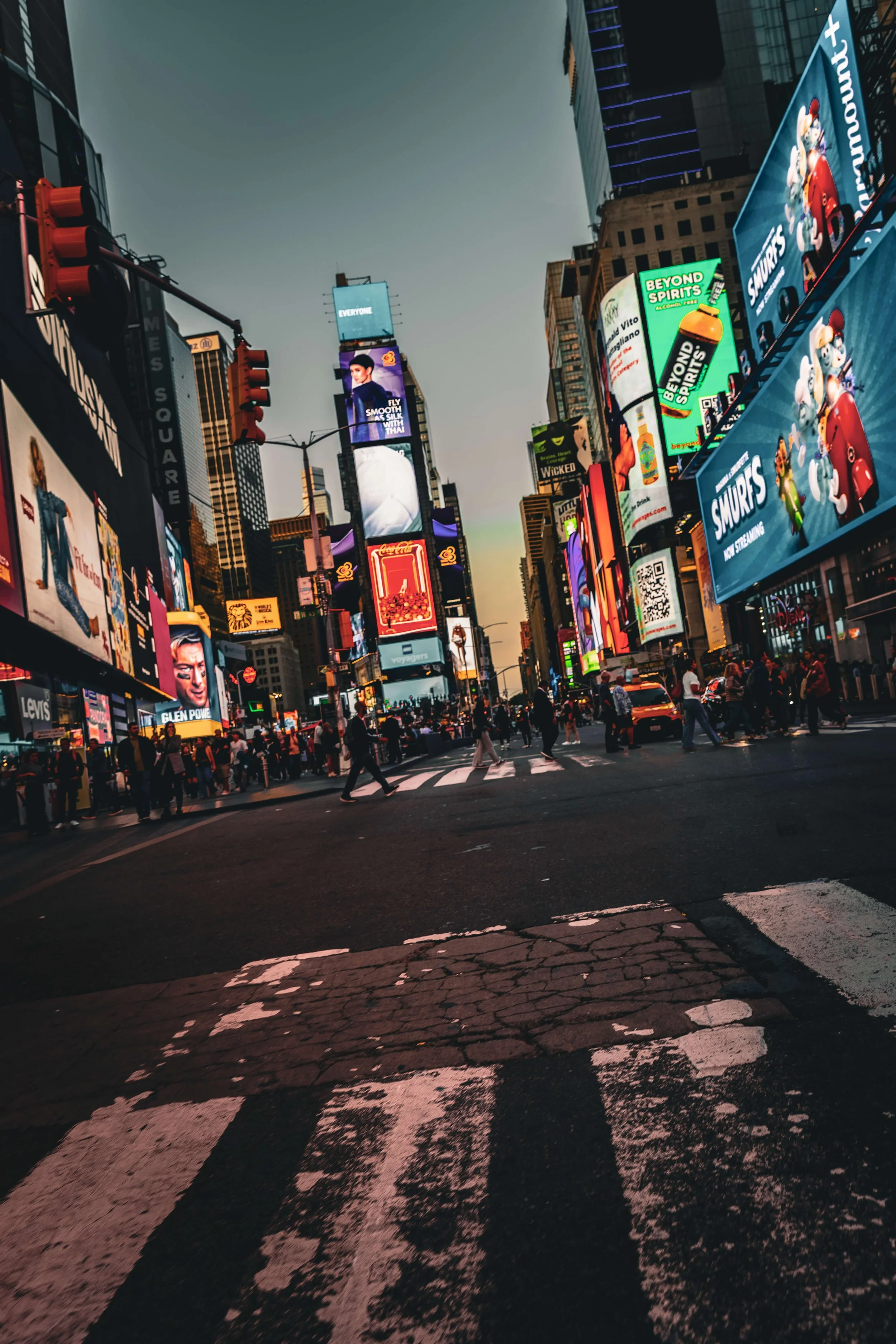 A bustling city intersection at dusk with bright electronic billboards, pedestrians crossing the street, and traffic waiting at a red light in Times Square, New York City.