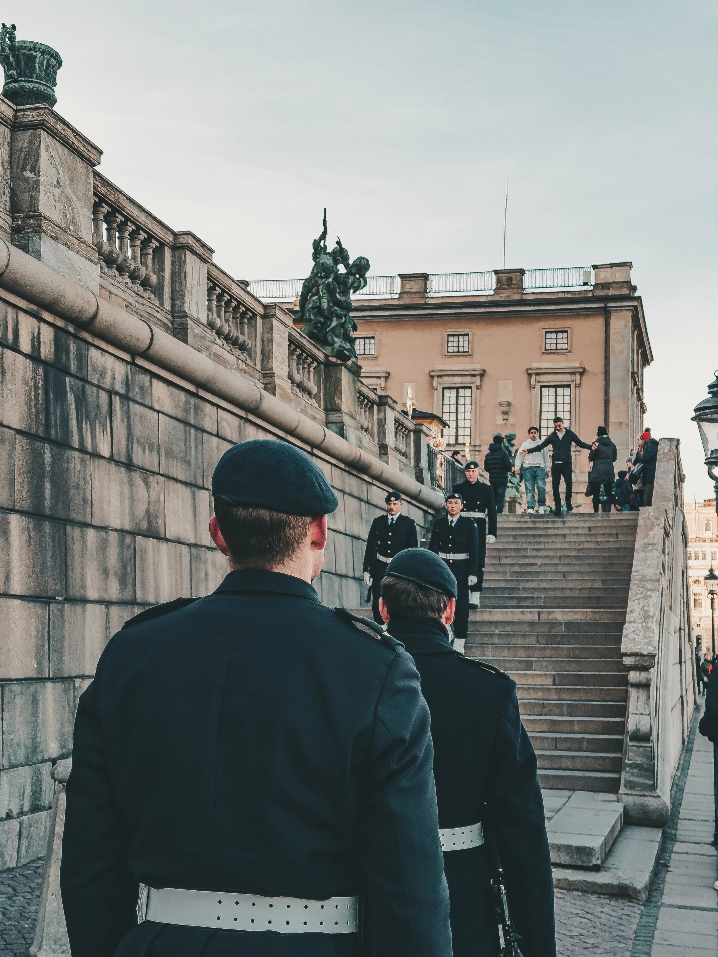 Two police officers in uniform standing at the bottom of a stone staircase on a city street, with several people walking up and down and others standing at the top of the stairs. Large historic building with windows and a statue on the ledge are visi