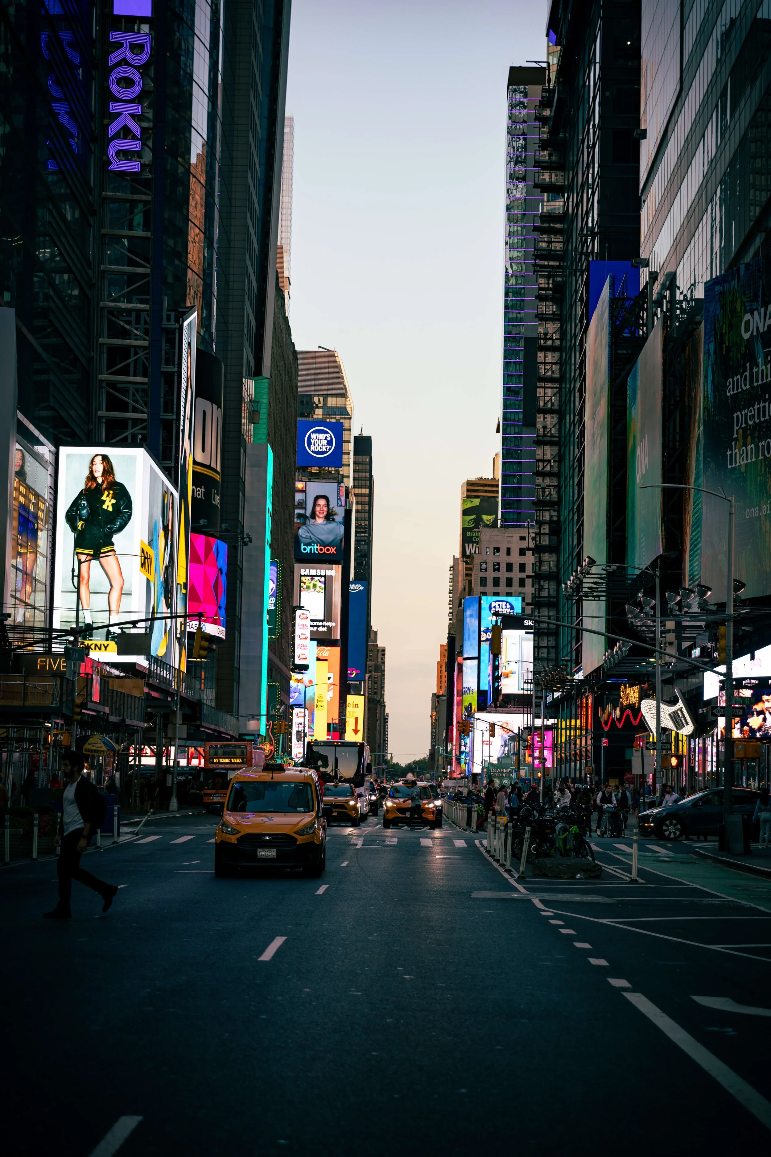 Times Square in New York City during dusk with tall buildings, bright electronic billboards, cars, taxis, and pedestrians.