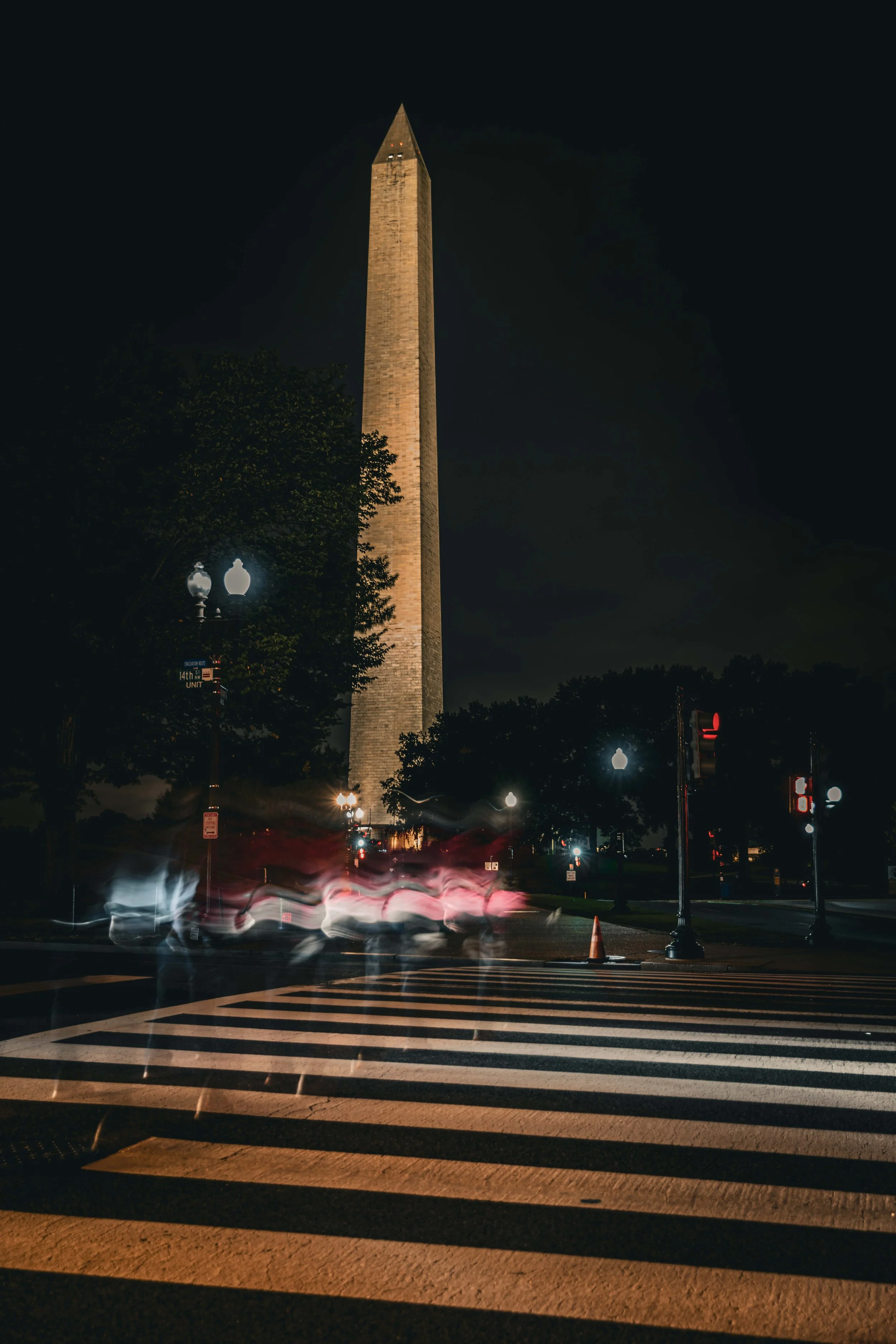 Nighttime view of the Washington Monument illuminated with streetlights and a crosswalk in the foreground, with blurry vehicle lights passing by.