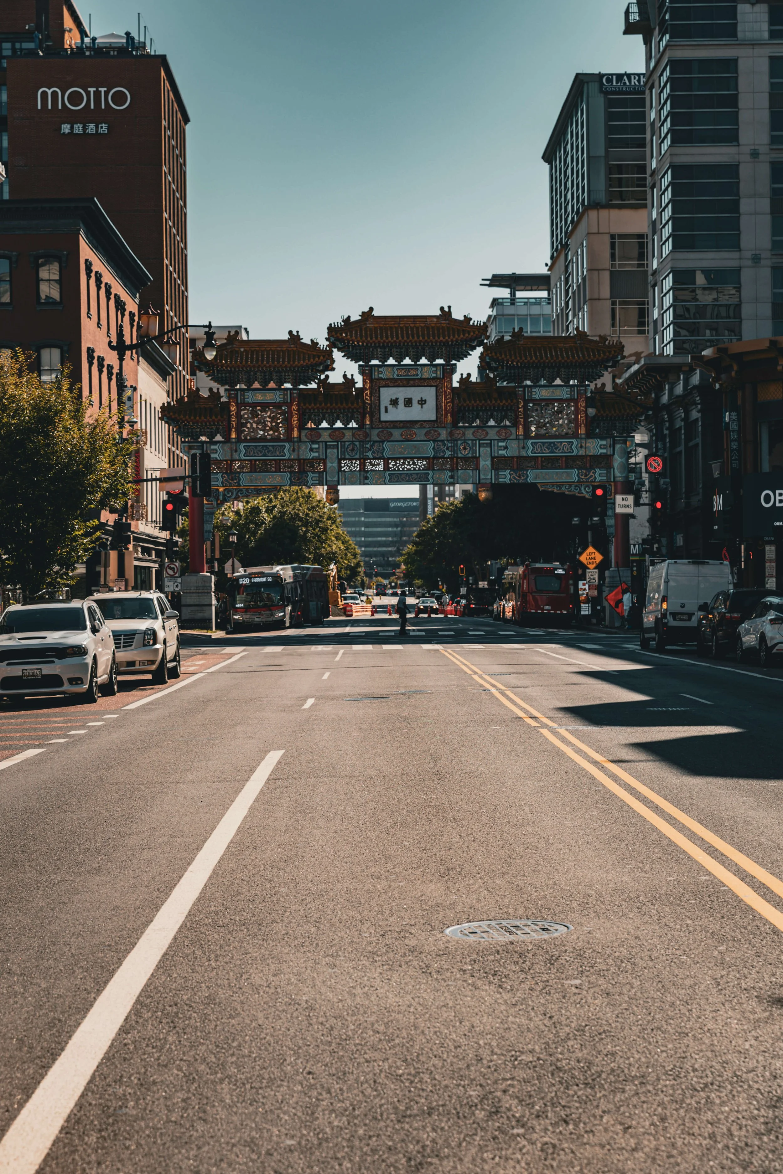 City street with Asian-style decorative gate spanning across the road, surrounded by modern buildings and parked cars under a clear sky.