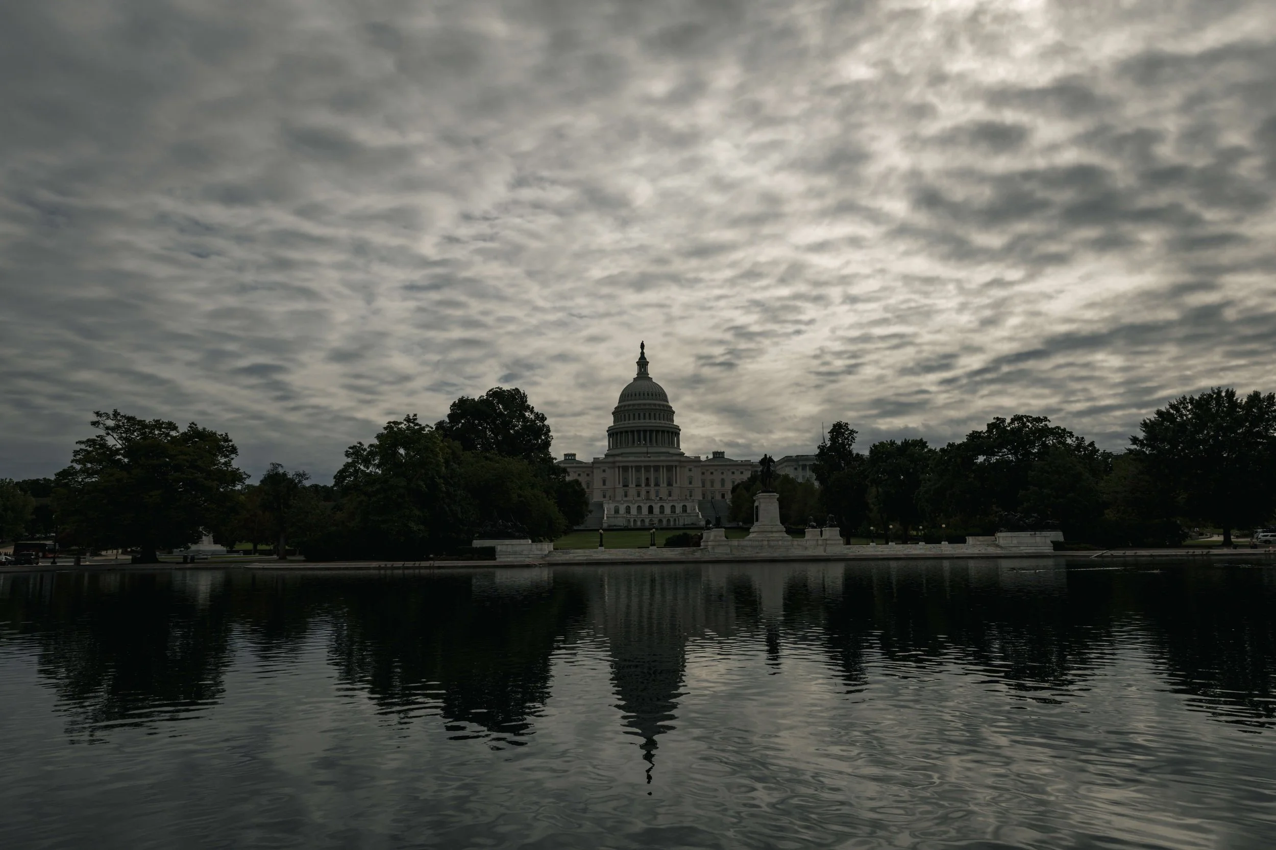 The Capitol building in Washington, D.C., with a reflection in a body of water, cloudy sky overhead.