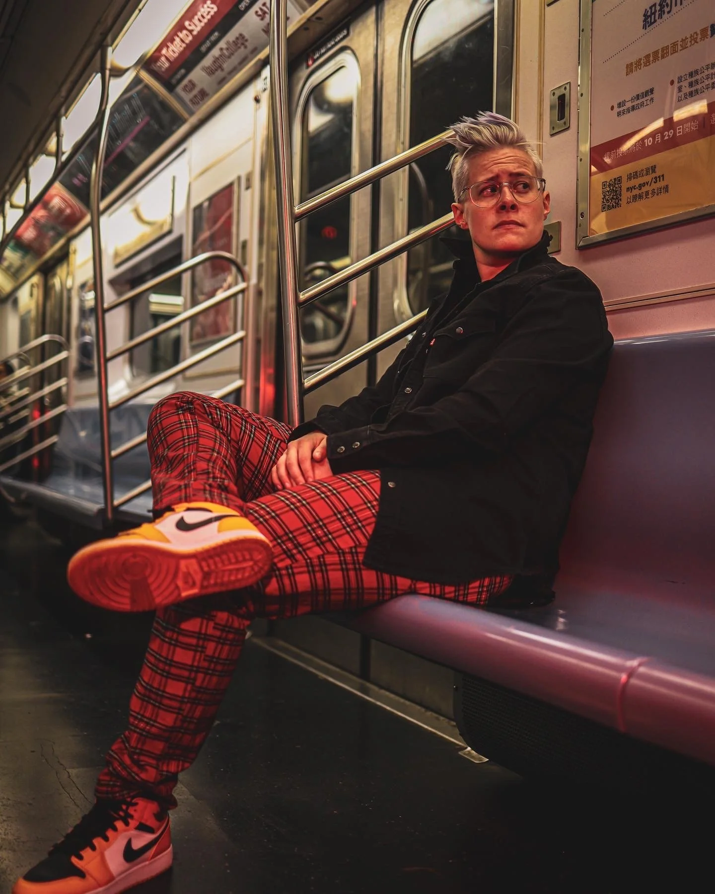 A young woman with glasses and styled silver hair sitting on a subway seat, wearing red plaid pants, black jacket, and Nike sneakers, looking off to the side in a subway car.