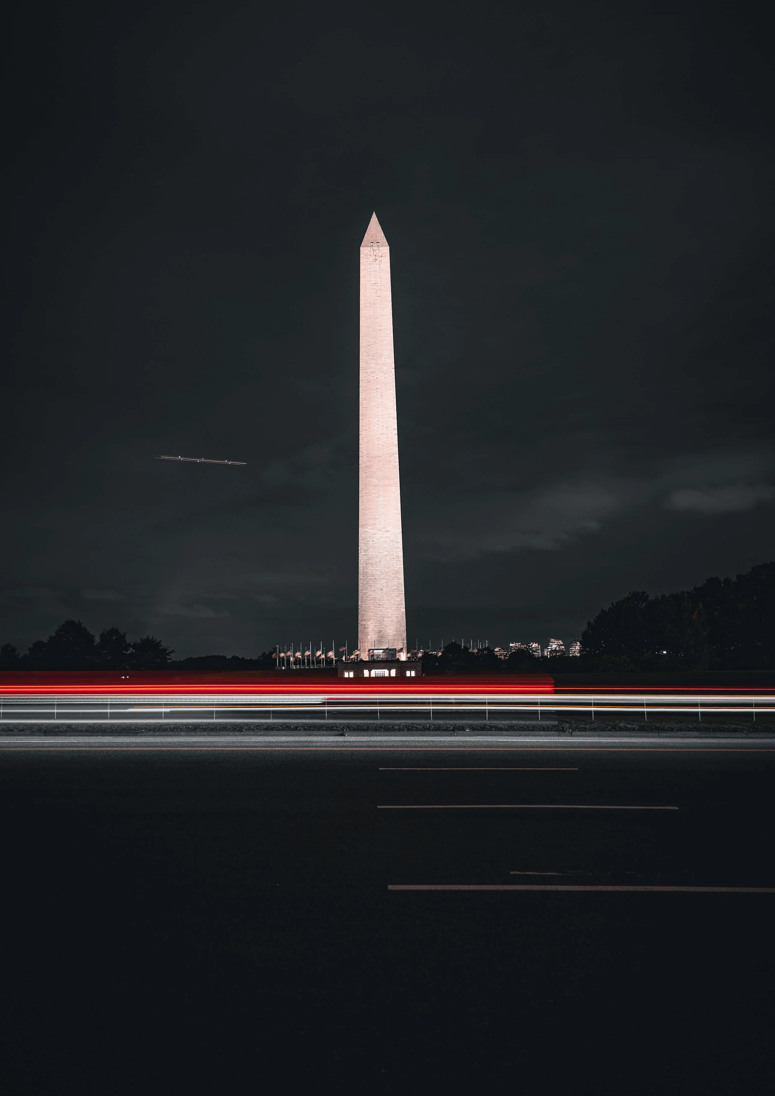 Nighttime view of the Washington Monument with light trails from passing vehicles on the road in the foreground.