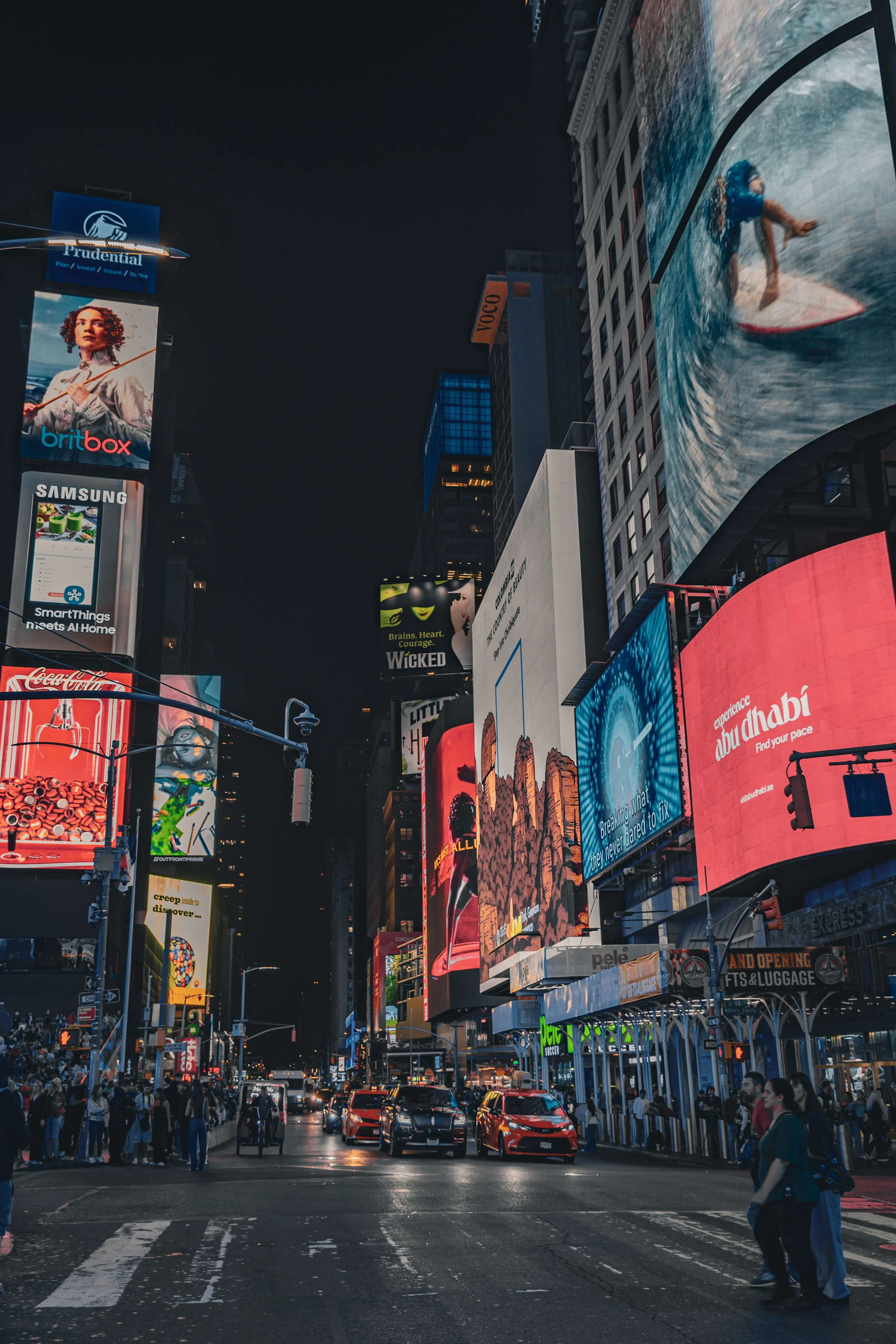 Brightly lit billboards and advertisements on buildings at night in Times Square, New York City, with cars and pedestrians in the foreground.
