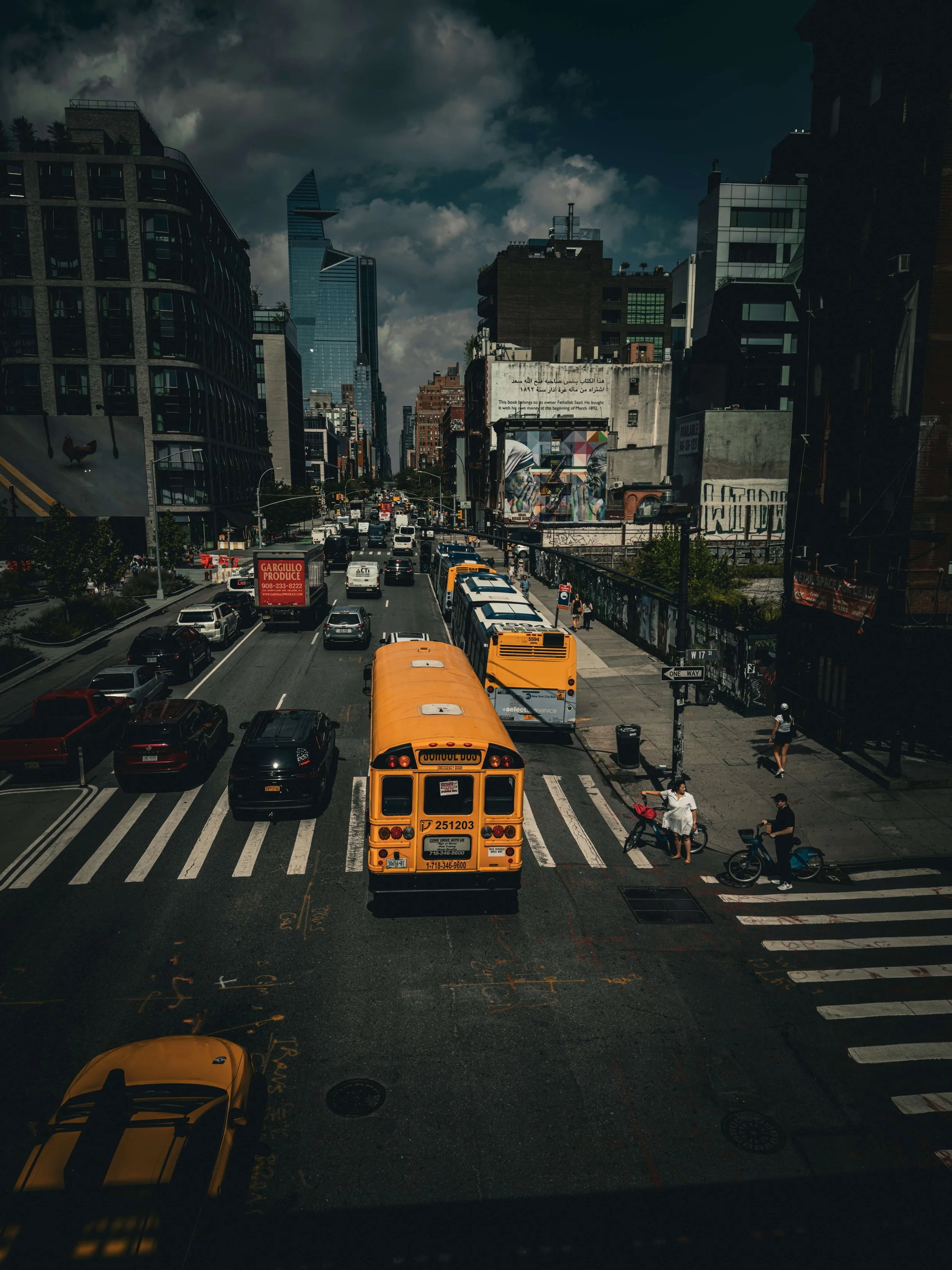 Urban street scene with yellow school buses, cars, pedestrians, and skyscrapers under a cloudy sky.