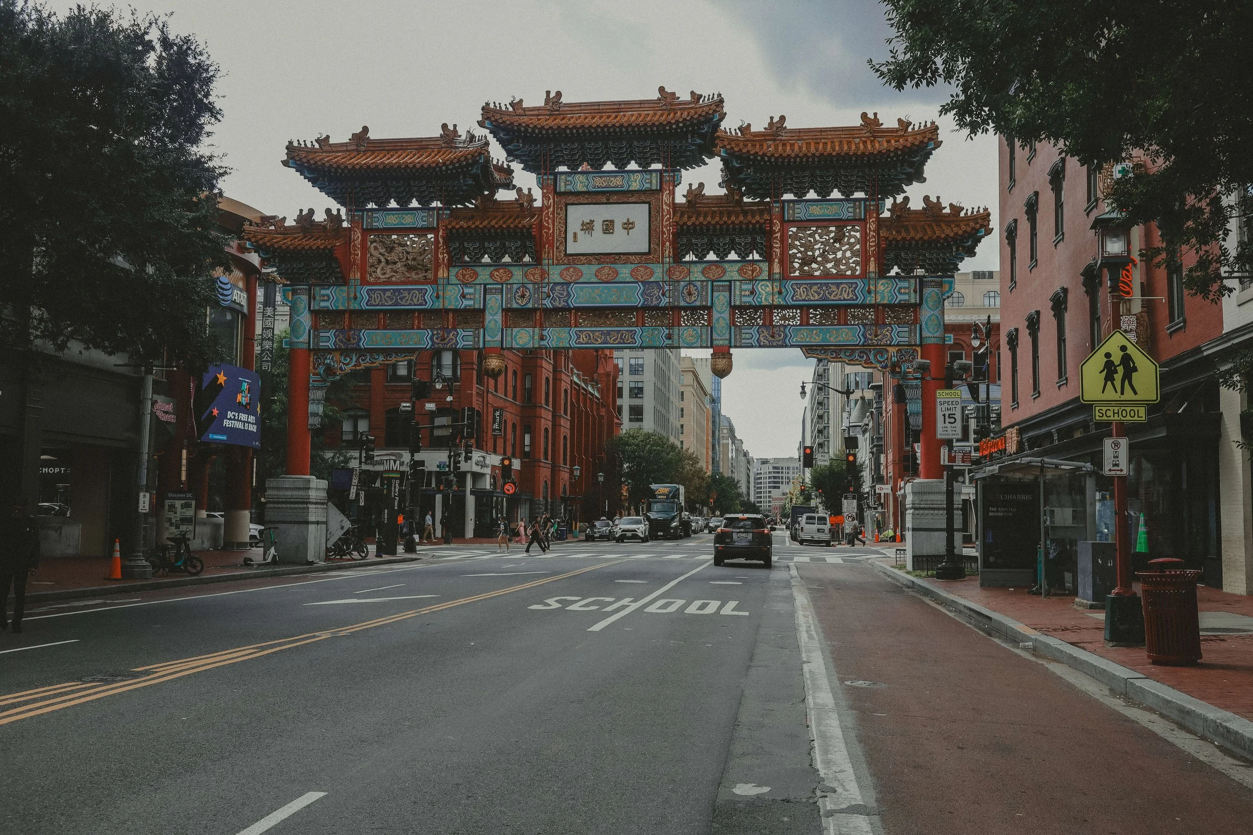 A city street with an ornate Chinese-style archway spanning across it. The street has cars, pedestrians, and buildings on either side. Signs indicate a school zone.