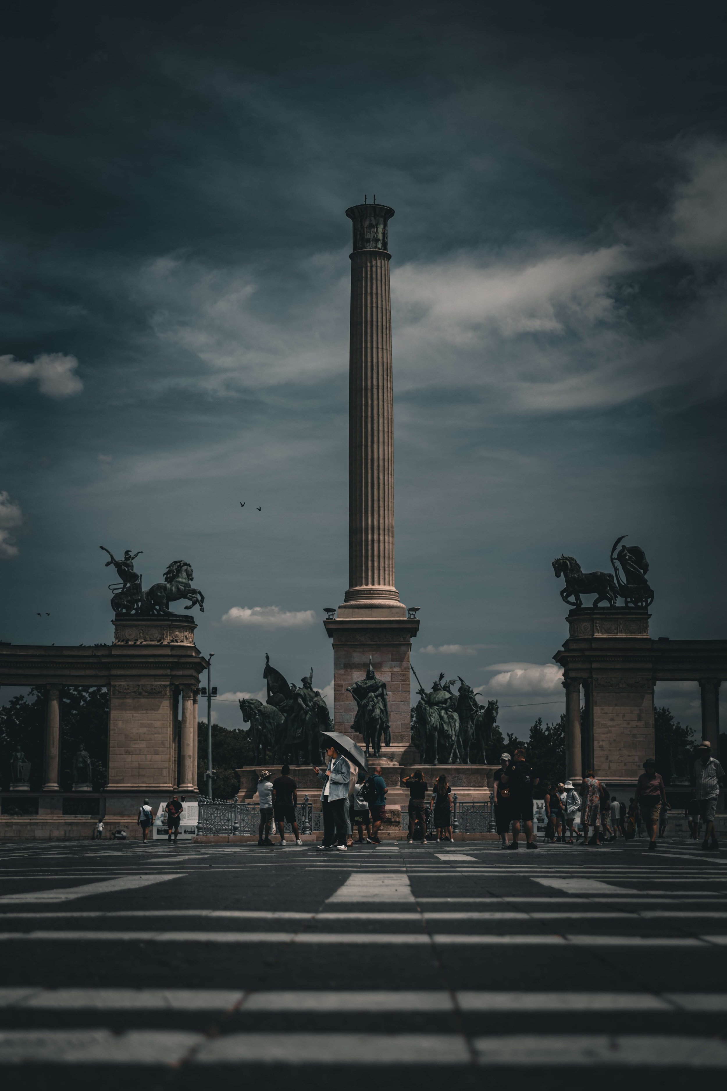 A large stone monument with a tall column topped with a rounded capital, surrounded by statues of horses and chariots at the base. There are people walking and standing in front of the monument, some holding umbrellas, with a dark, cloudy sky overhea
