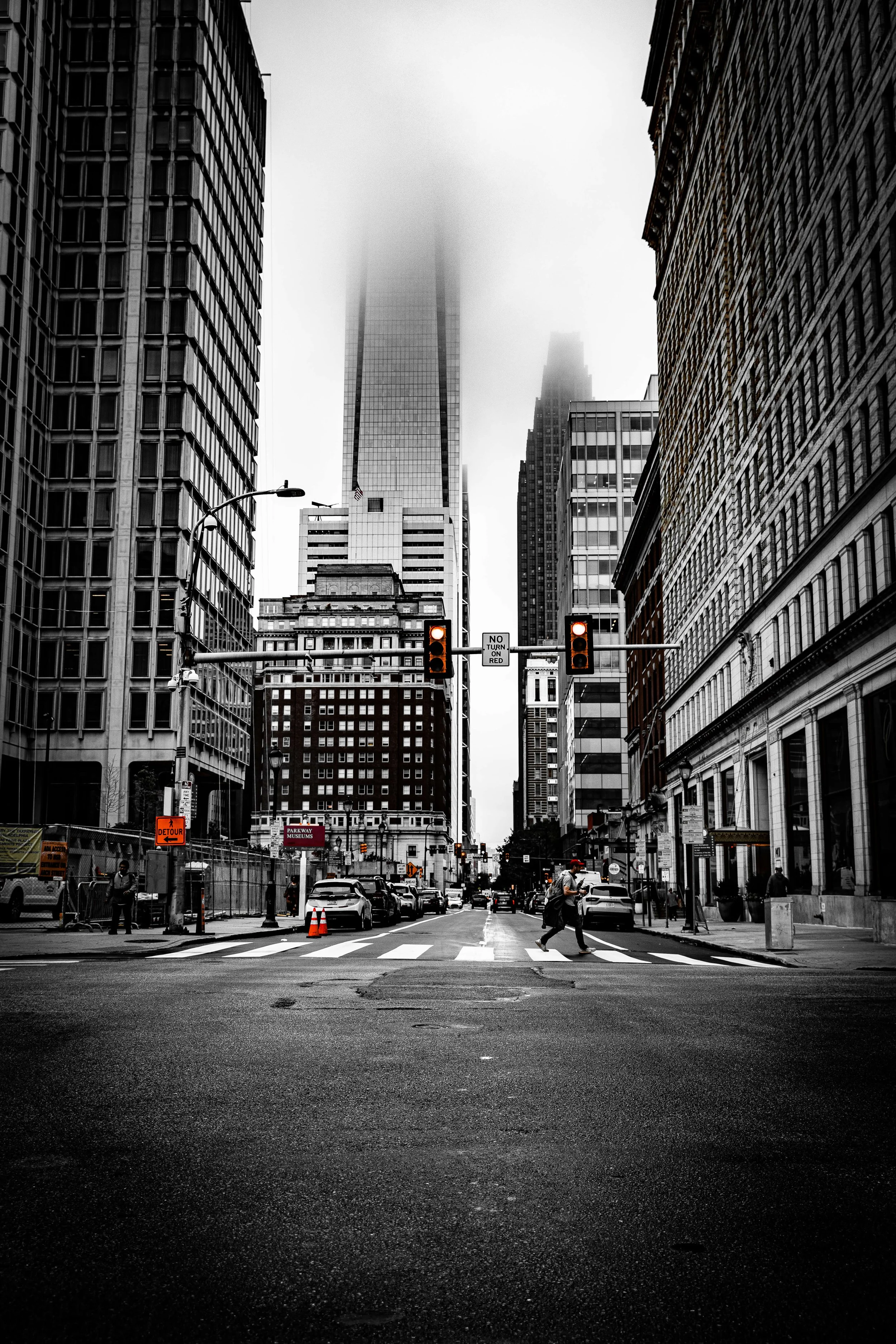 Black and white photo of a city street with tall skyscrapers, some obscured by fog, and a few pedestrians crossing at a crosswalk.