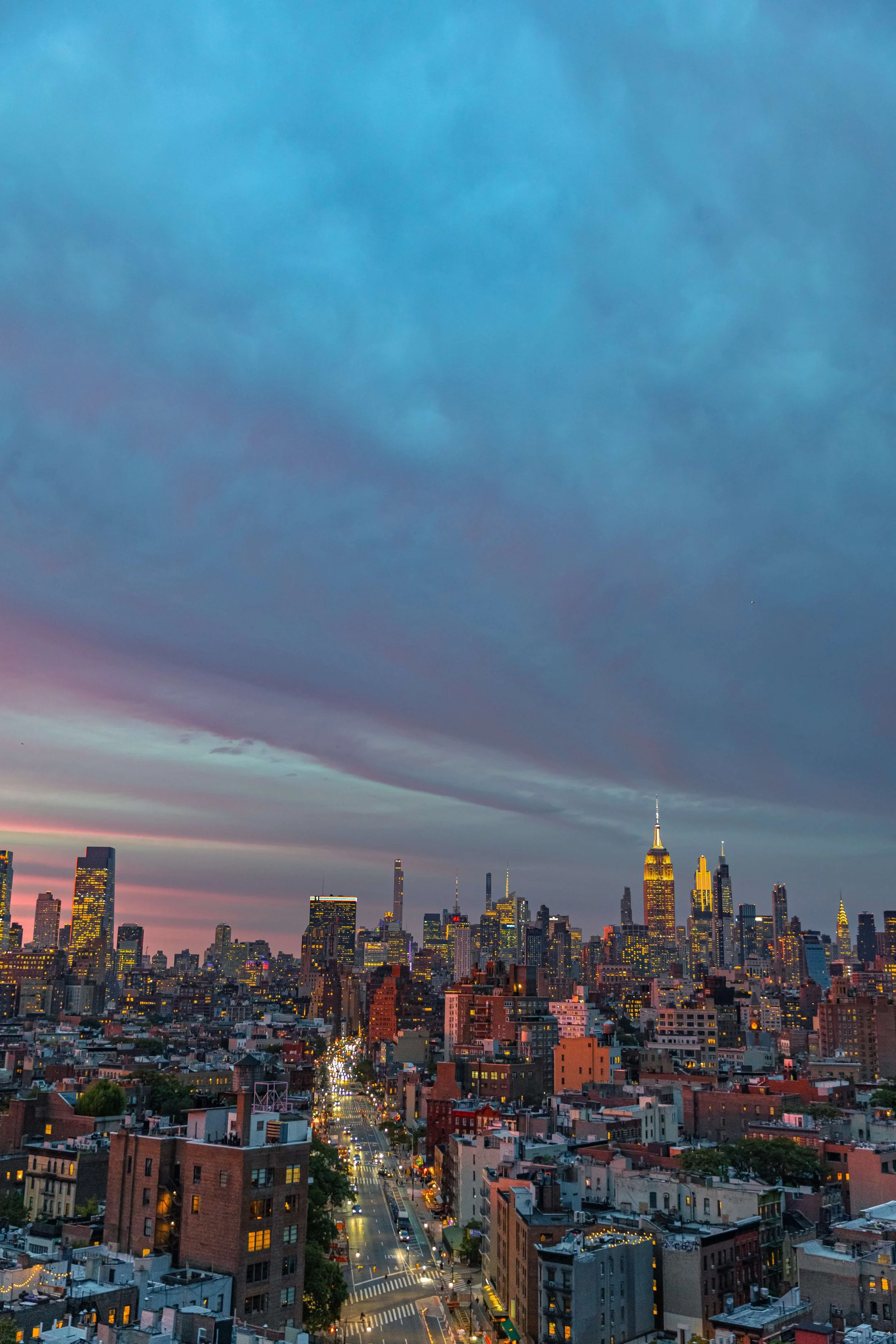A cityscape of New York City at dusk, with illuminated buildings and a sky with dark clouds and a hint of sunset colors.