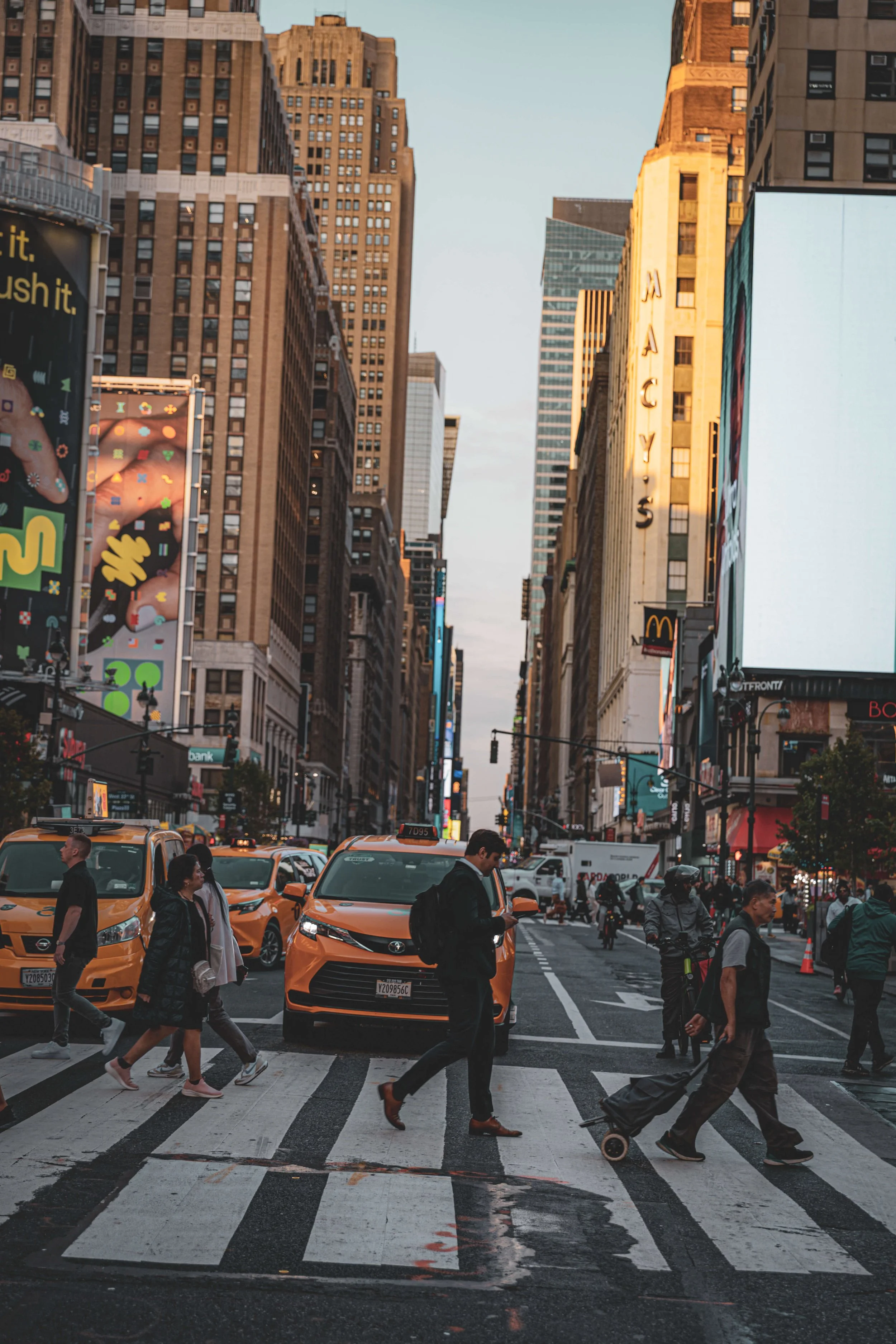 A busy city intersection with pedestrians crossing the street, yellow taxis, and tall buildings, including a Macy's sign, during sunset.