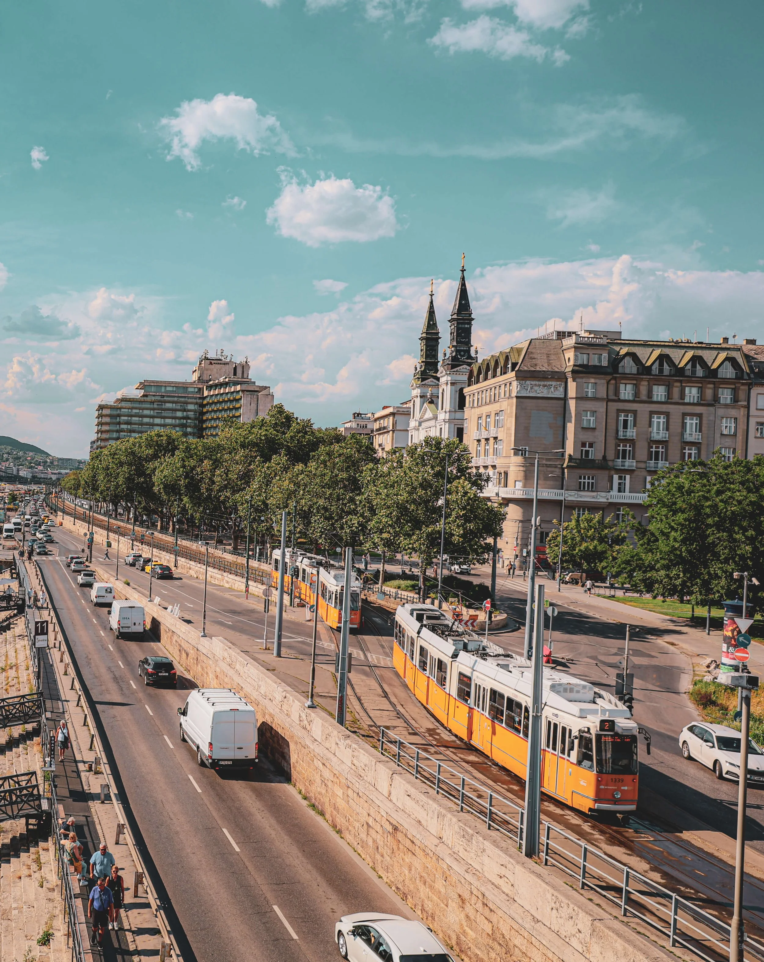 A cityscape showing a major road with cars and pedestrians, a tram on tracks with orange and white colors, and buildings including a church with twin spires, under a partly cloudy sky.