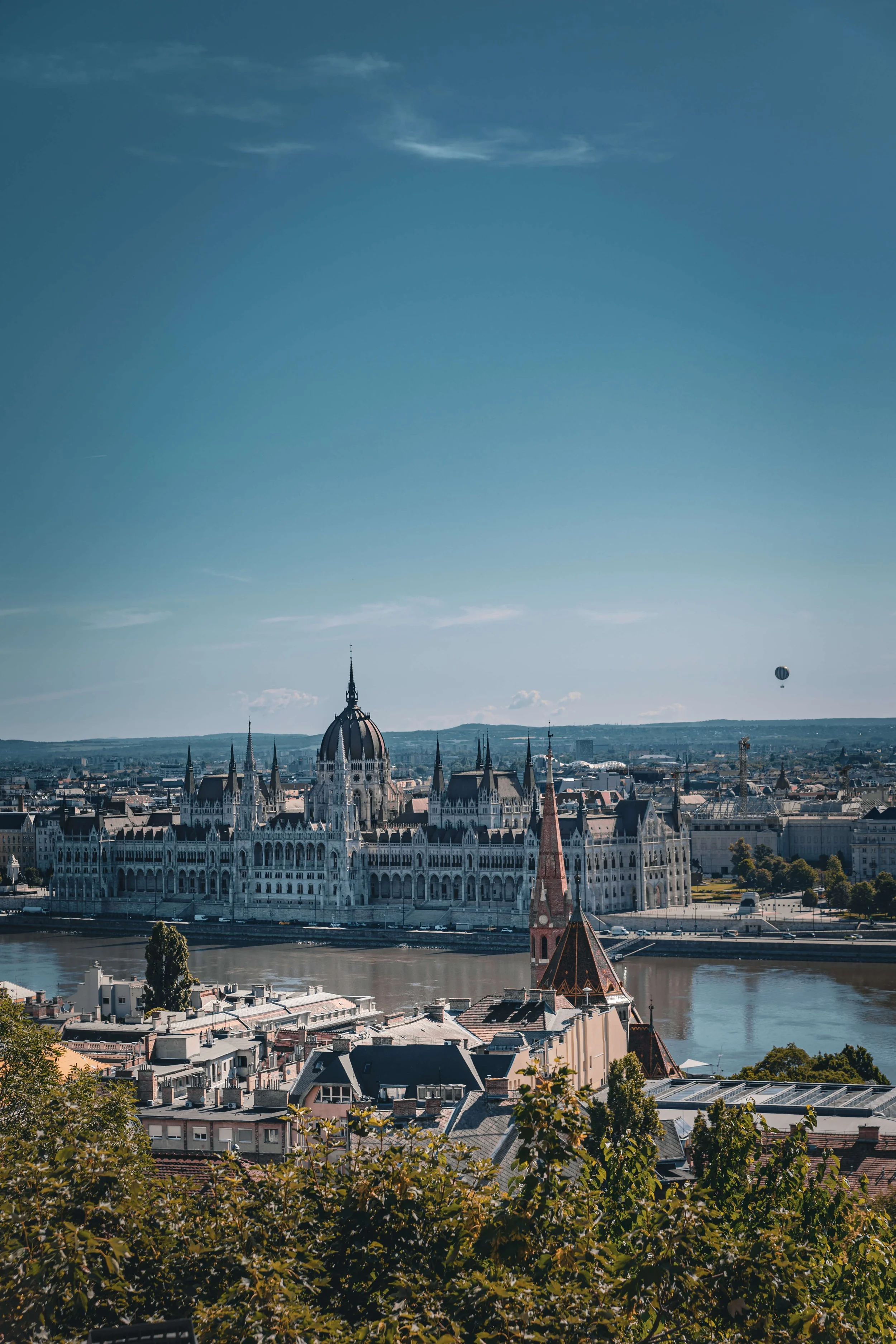 A scenic cityscape view featuring a historic building with ornate architecture, a river, and a church with a steeple. A hot air balloon floats in a clear blue sky above the city.