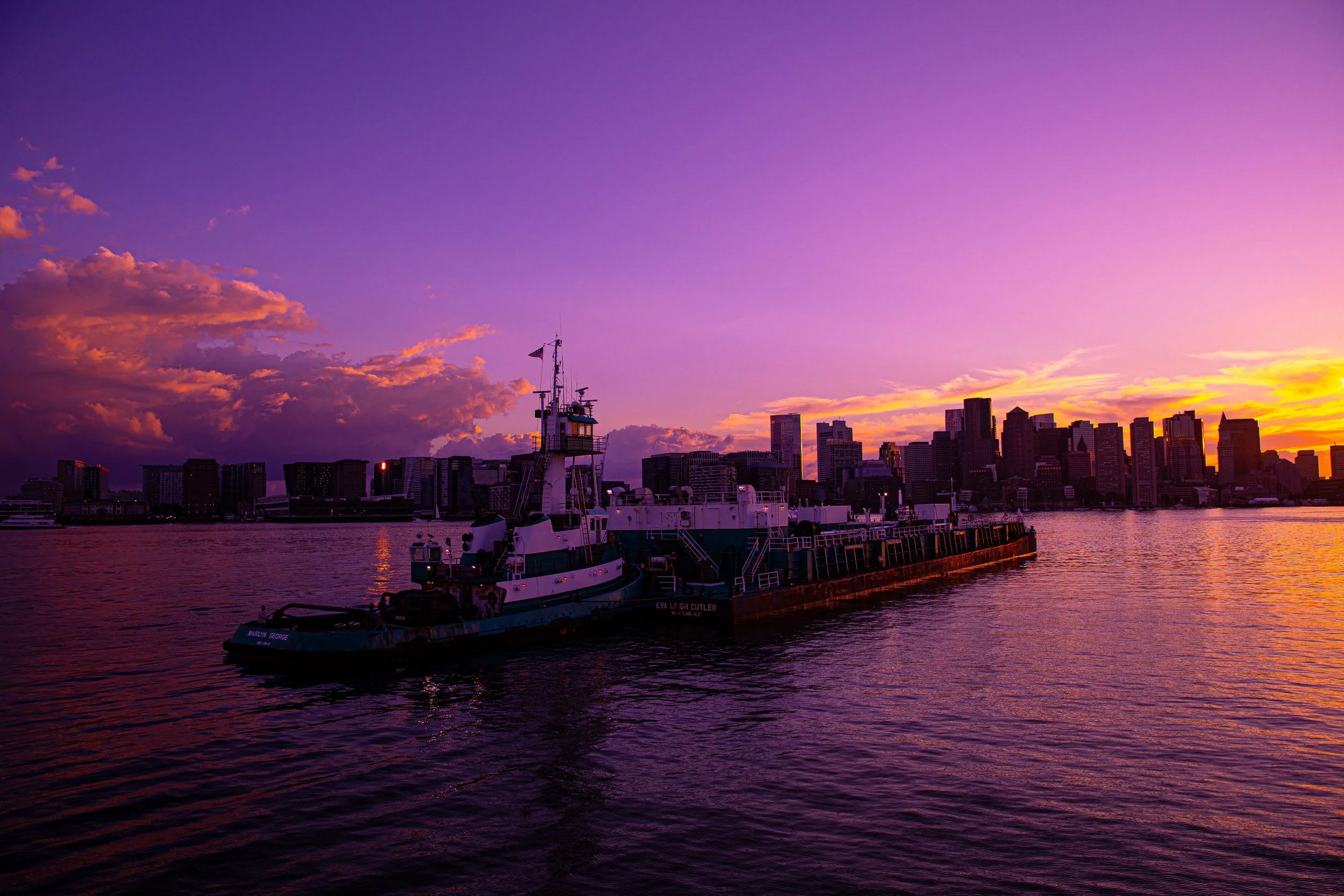 A boat on a body of water during sunset with a city skyline in the background and colorful sky with purple, pink, and orange hues.