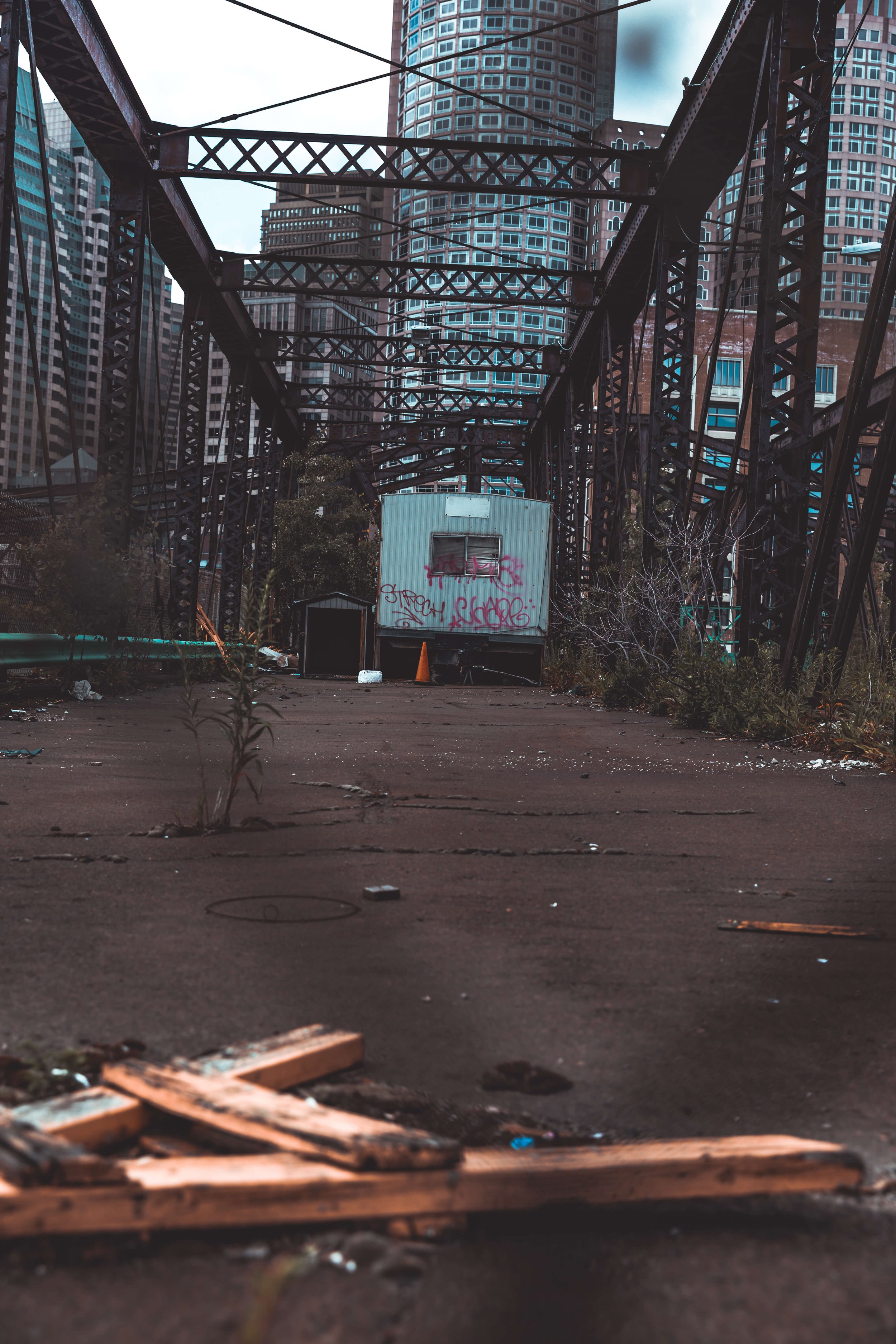 An abandoned lot with overgrown weeds and debris, surrounded by tangled metal structures; in the background, tall skyscrapers dominate the skyline.