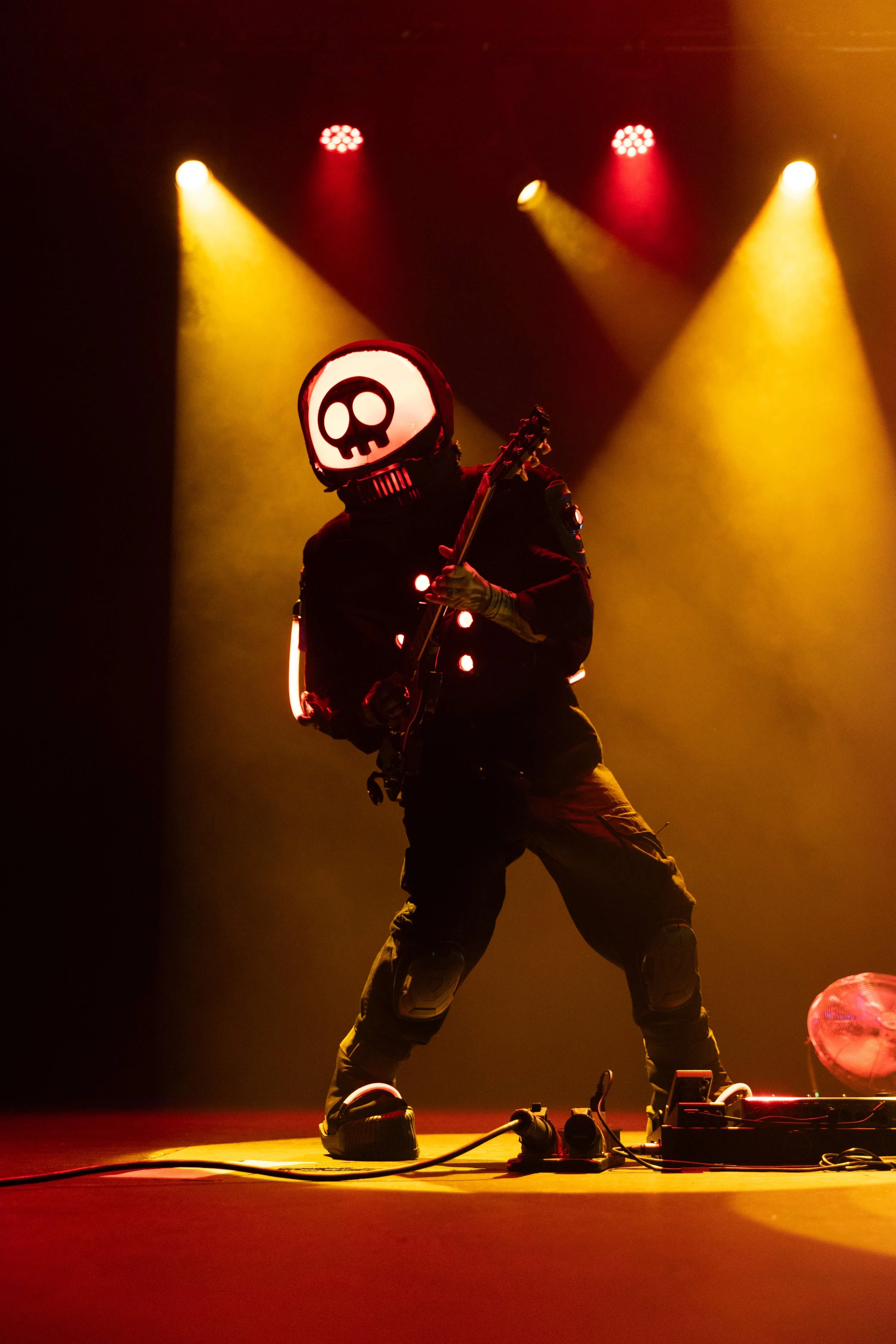 A person wearing a helmet with a skull face design, playing an electric guitar on stage with colorful spotlights.