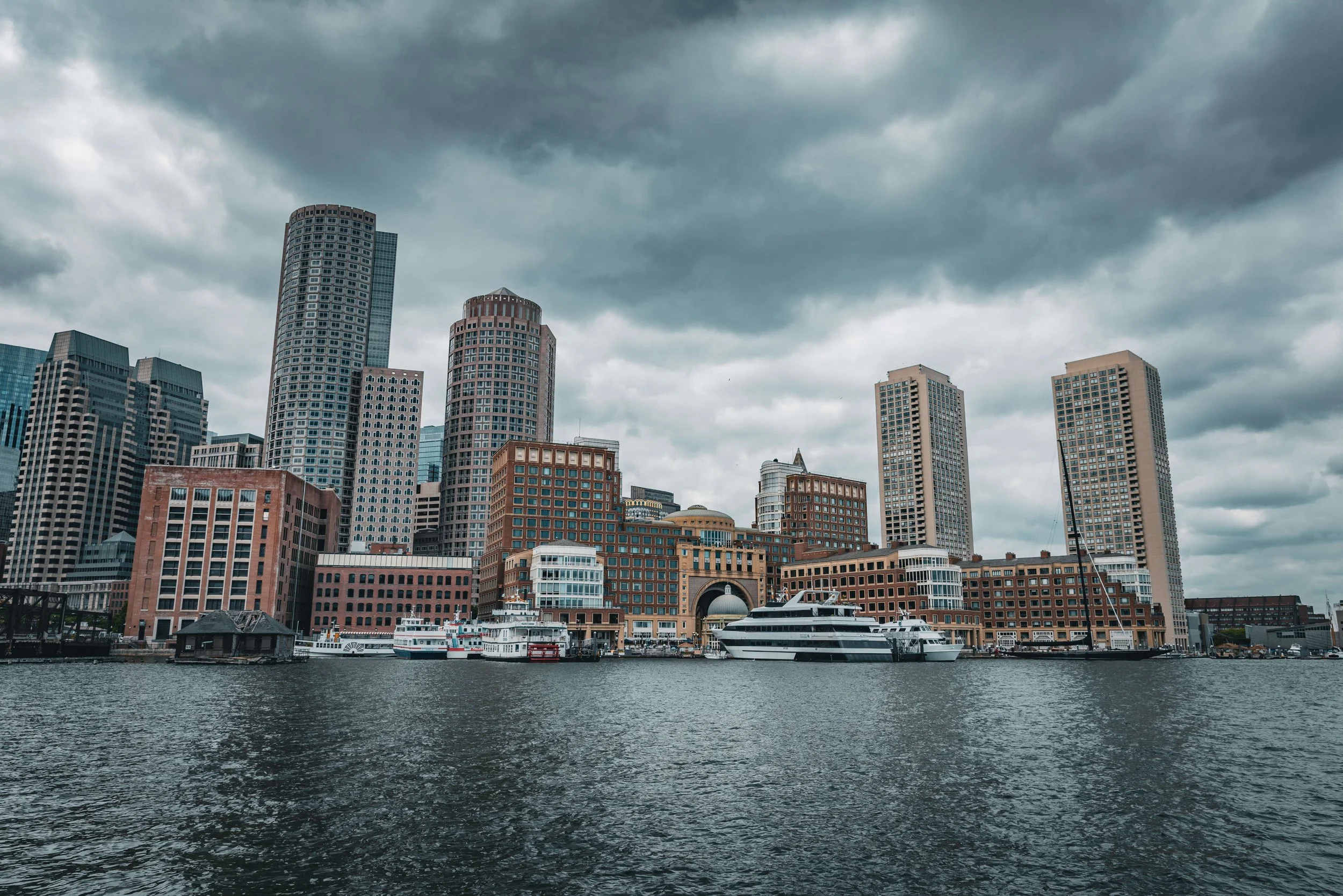 A city skyline featuring tall skyscrapers with a waterfront and boats docked along the water's edge, under a cloudy sky.