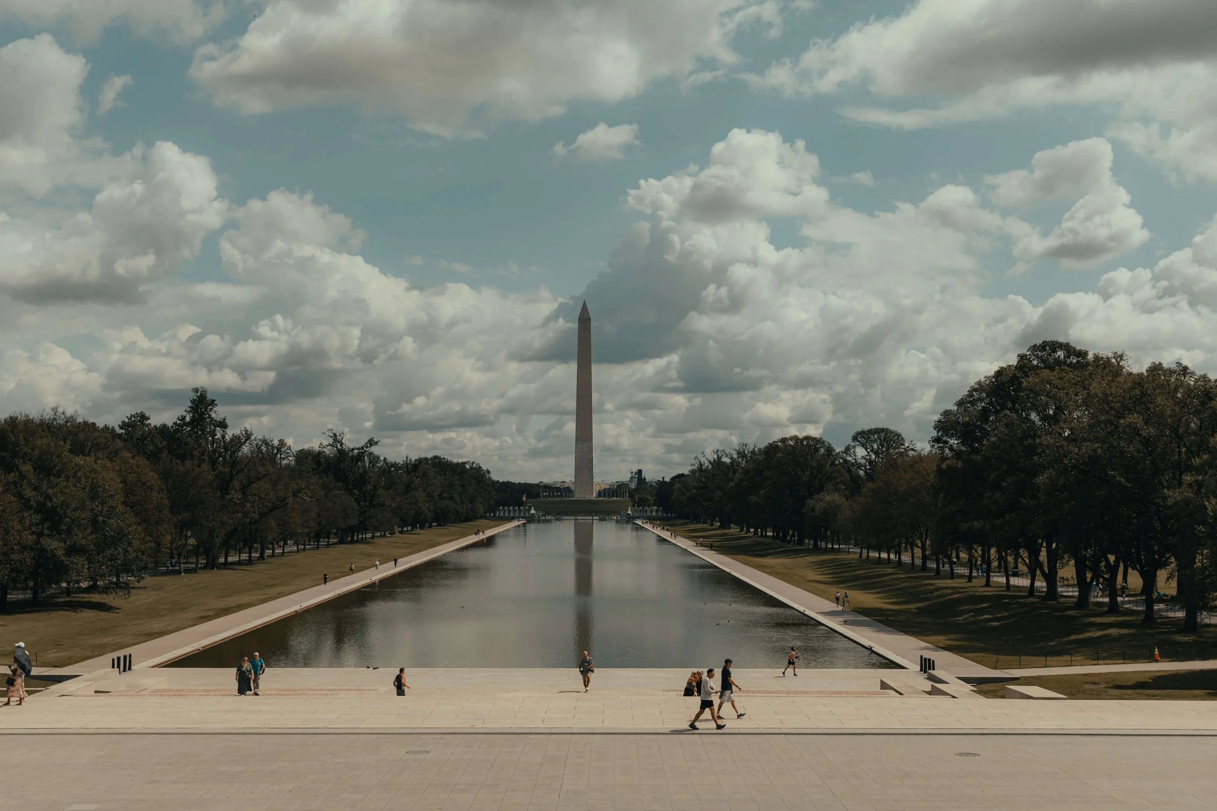 Washington Monument viewed from the Reflecting Pool on a partly cloudy day with people walking along the pool and the steps in the foreground.