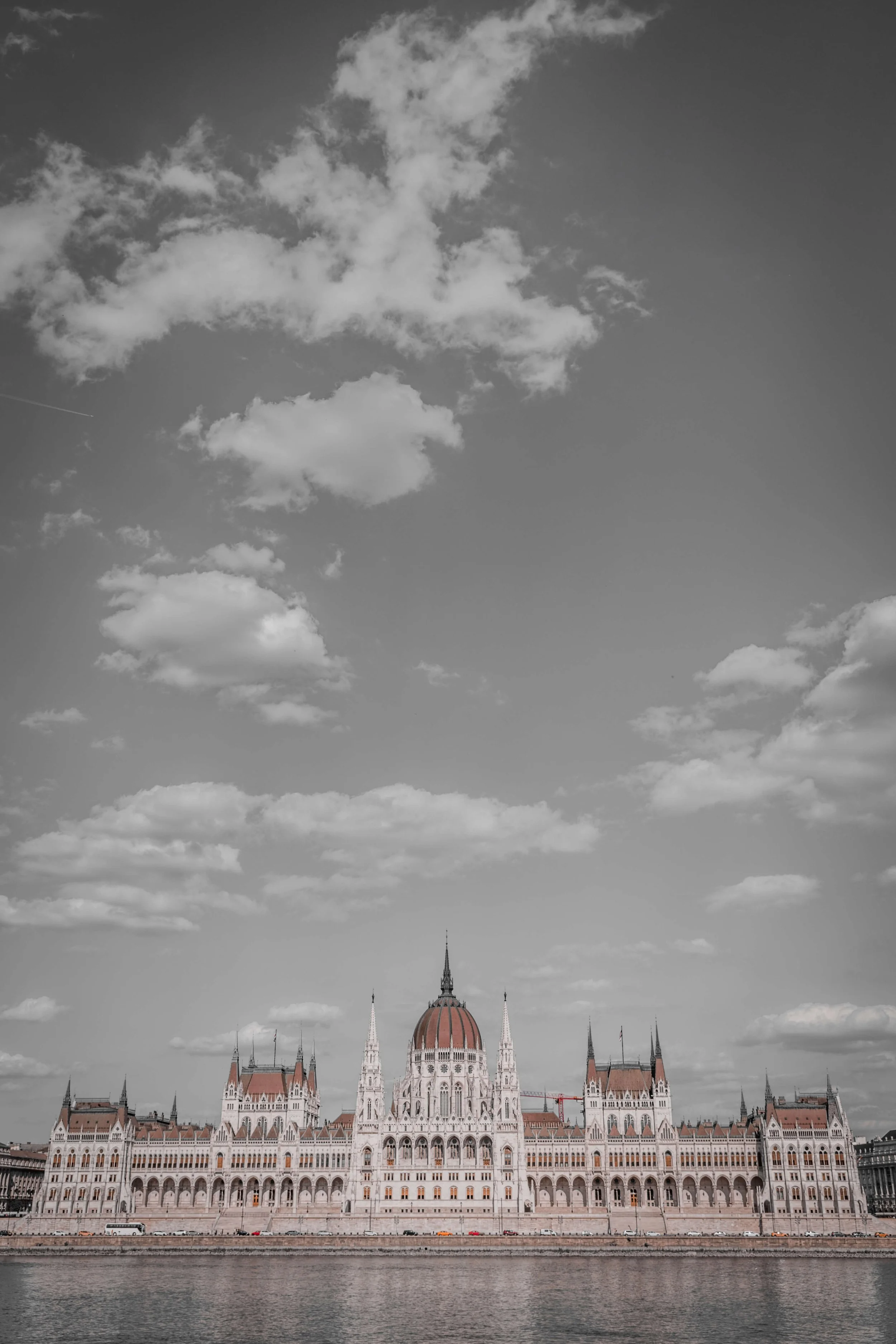Black and white photo of the Hungarian Parliament building reflected in the water, with scattered clouds in the sky.
