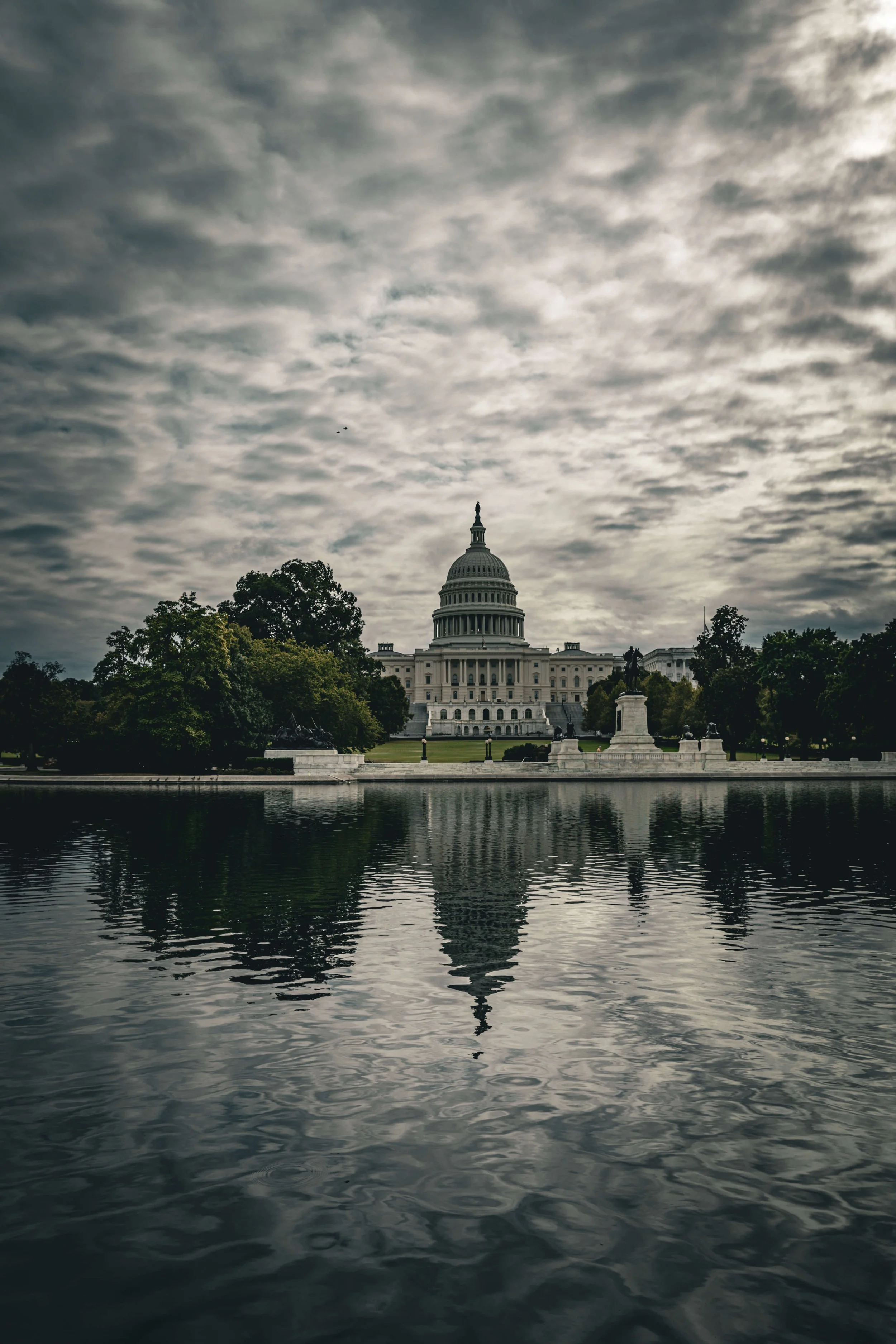 View of the United States Capitol building reflecting on the water with a cloudy sky overhead.