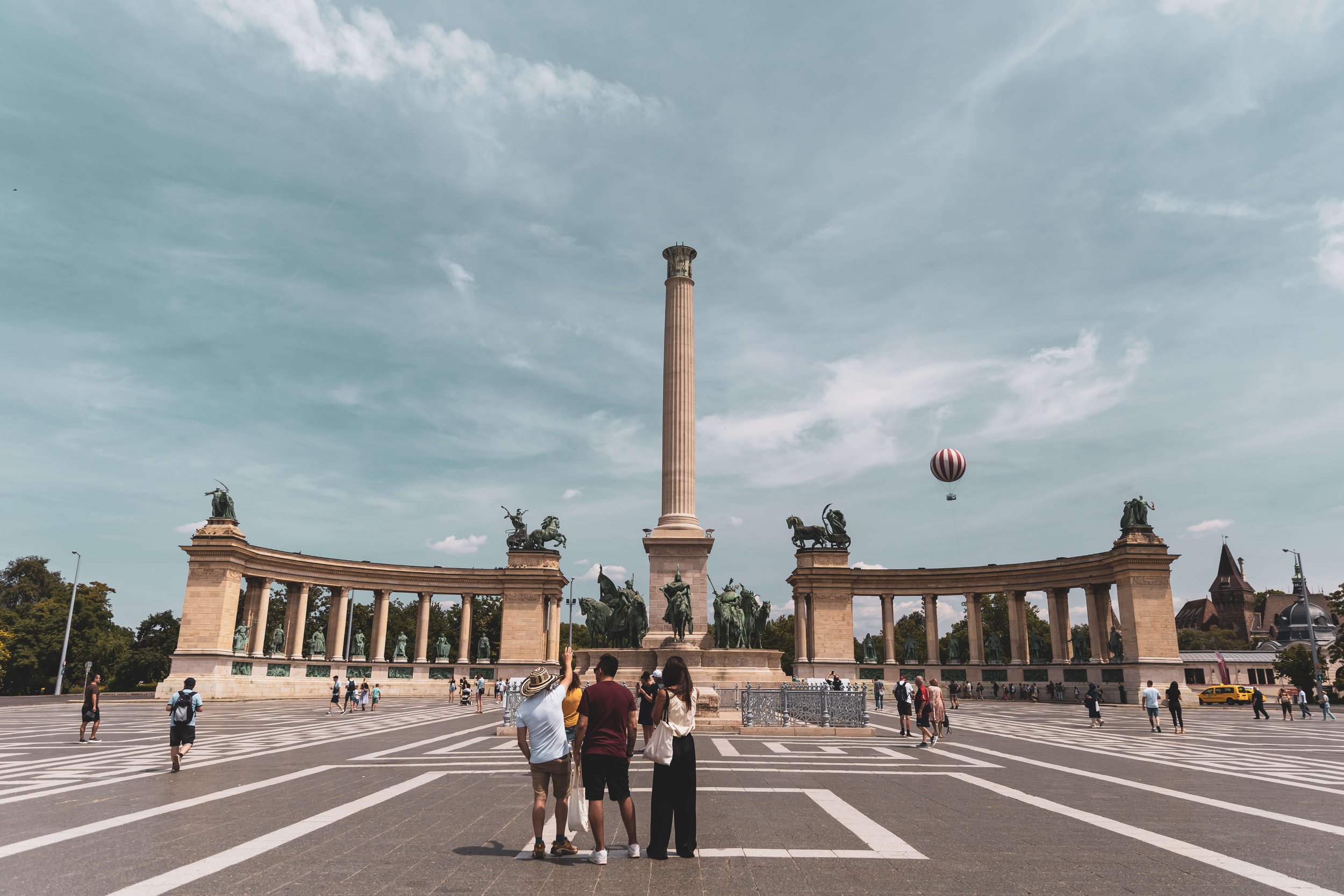 View of Millennium Monument in Heroes' Square Budapest, with statues of Hungarian national leaders, a tall column, and several tourists taking photos under a partly cloudy sky.