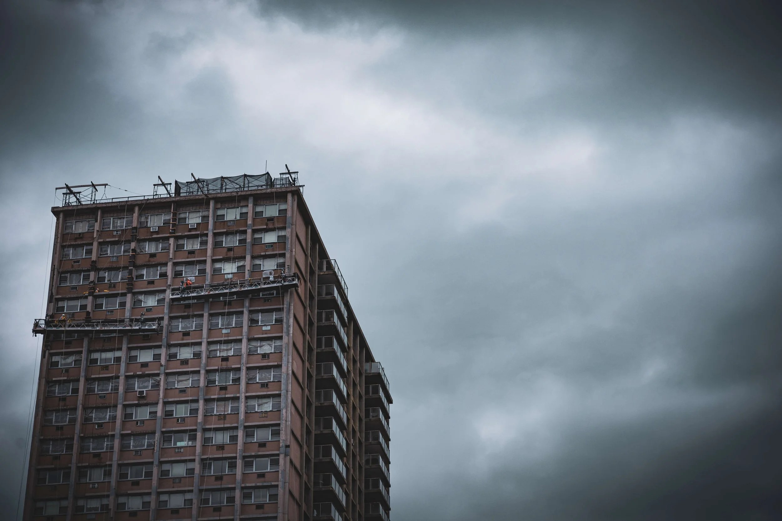 A high-rise apartment building under dark, cloudy skies with window air conditioning units and exterior maintenance work in progress.