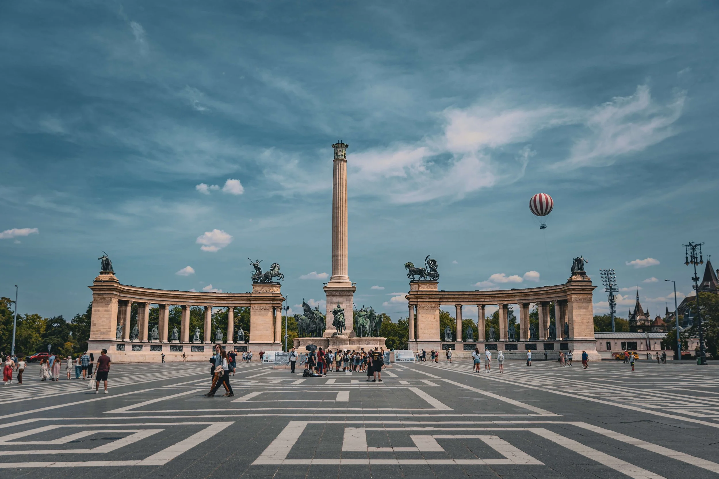 A large public square with a monument featuring a tall column topped with a statue, surrounded by sculptures of horses and chariots, and an archway with columns. There are people walking and gathering in the square on a sunny day with a blue sky and 