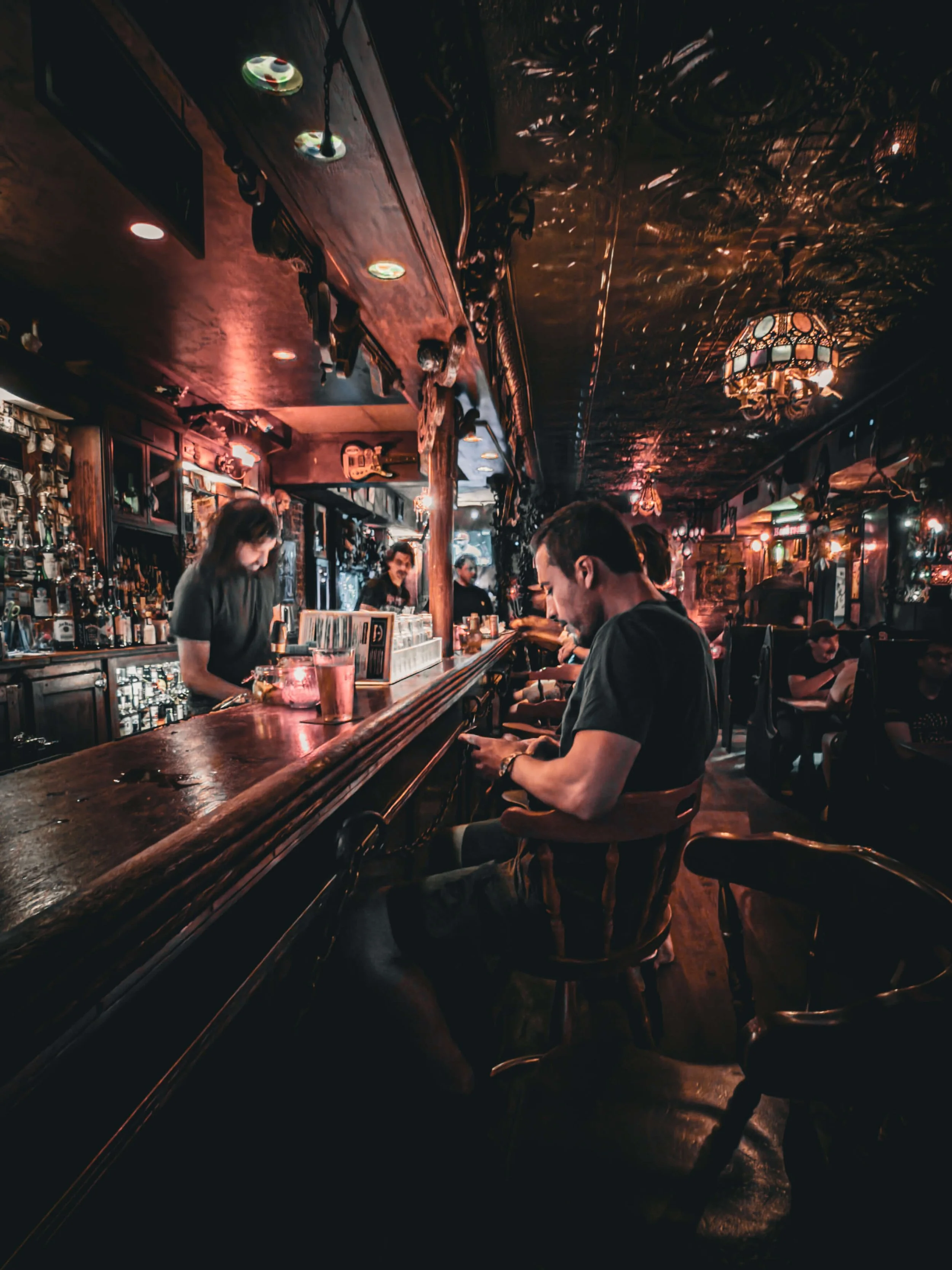 People sitting at a bar, with a bartender preparing drinks, in a dimly lit, cozy pub with wooden decor and vintage lighting.