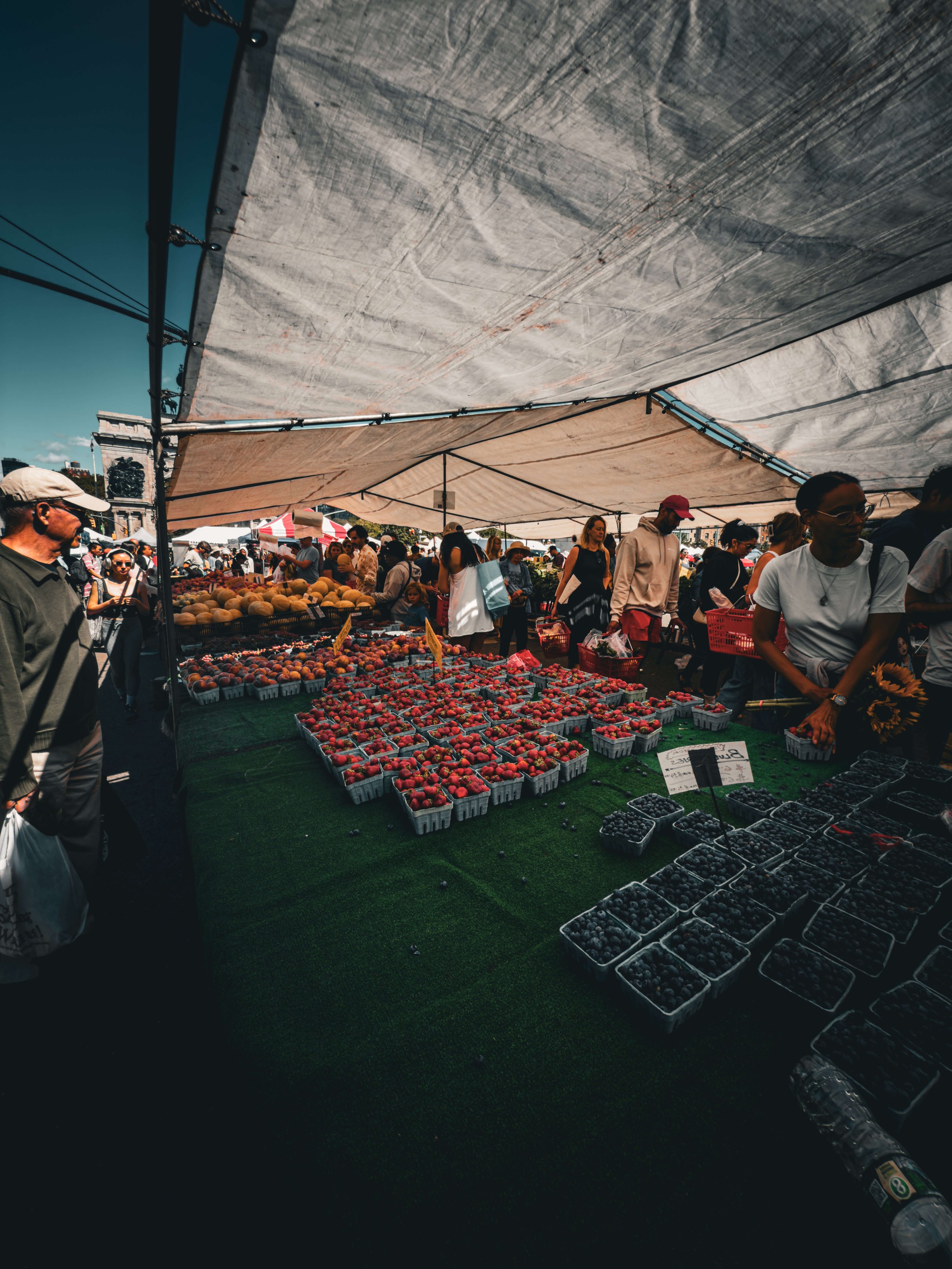 People shopping at an outdoor farmers market stall with strawberries, blueberries, and other fresh berries under a large canopy.