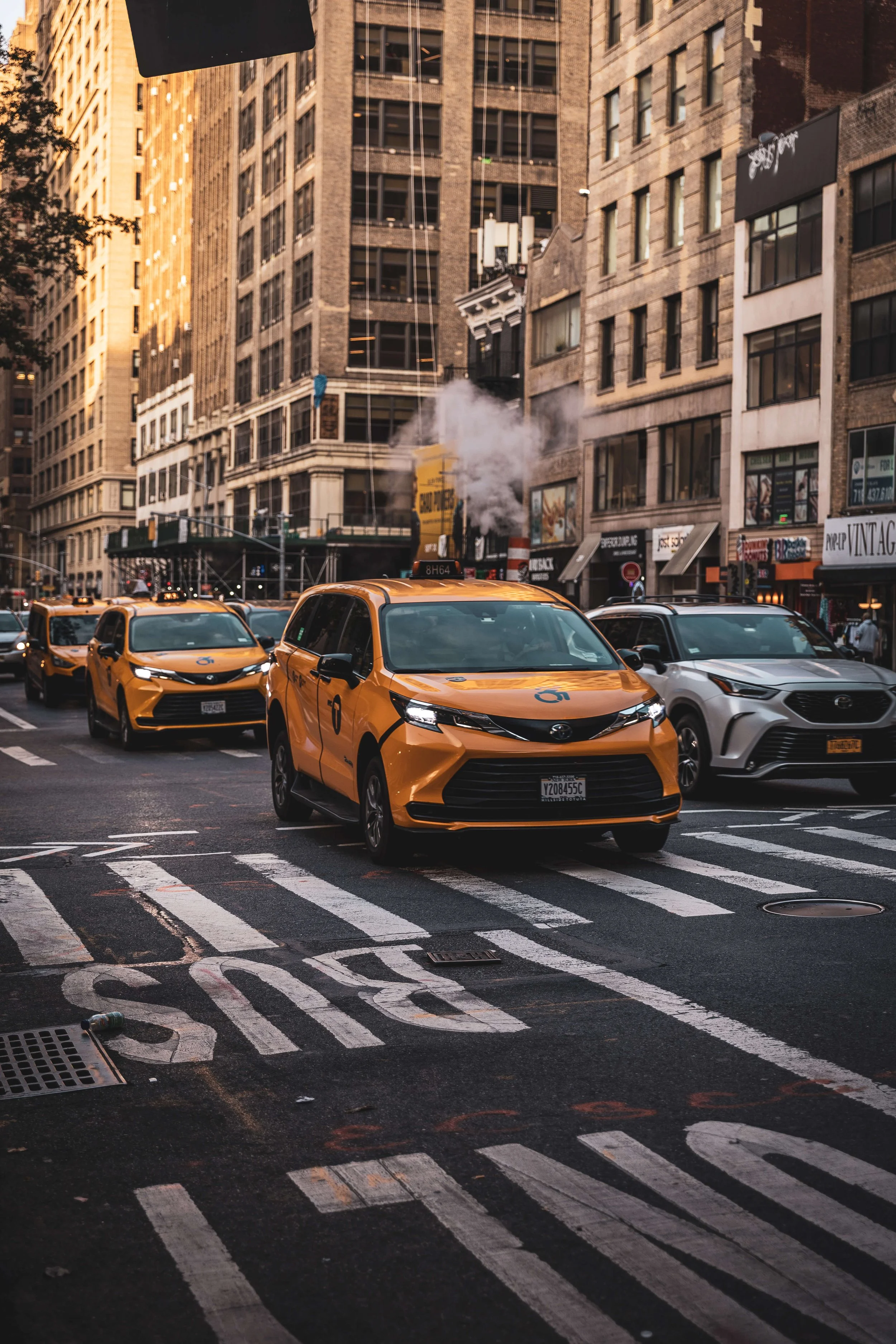 A city street with yellow taxis and a silver car at a crosswalk, tall buildings on both sides, and a cloud of steam rising from a manhole.