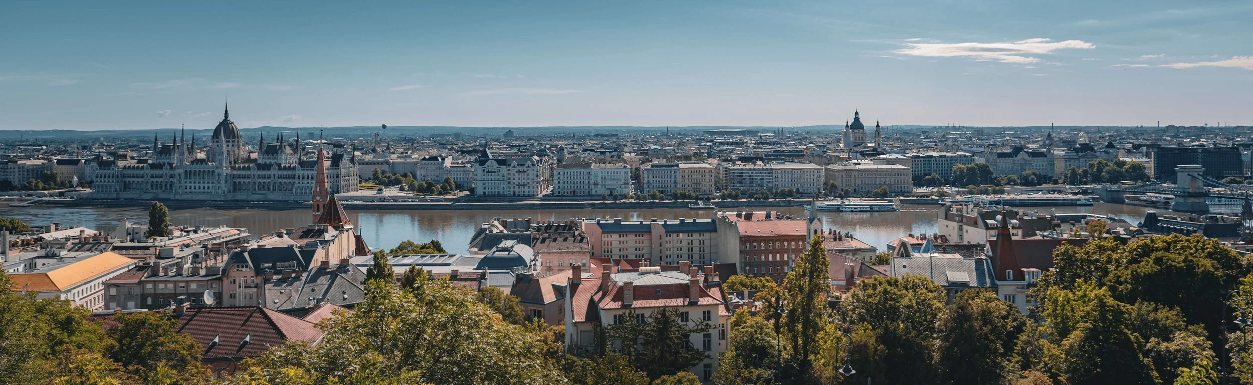 A panoramic daytime view of Budapest, Hungary, featuring the Parliament Building along the Danube River and the Chain Bridge.