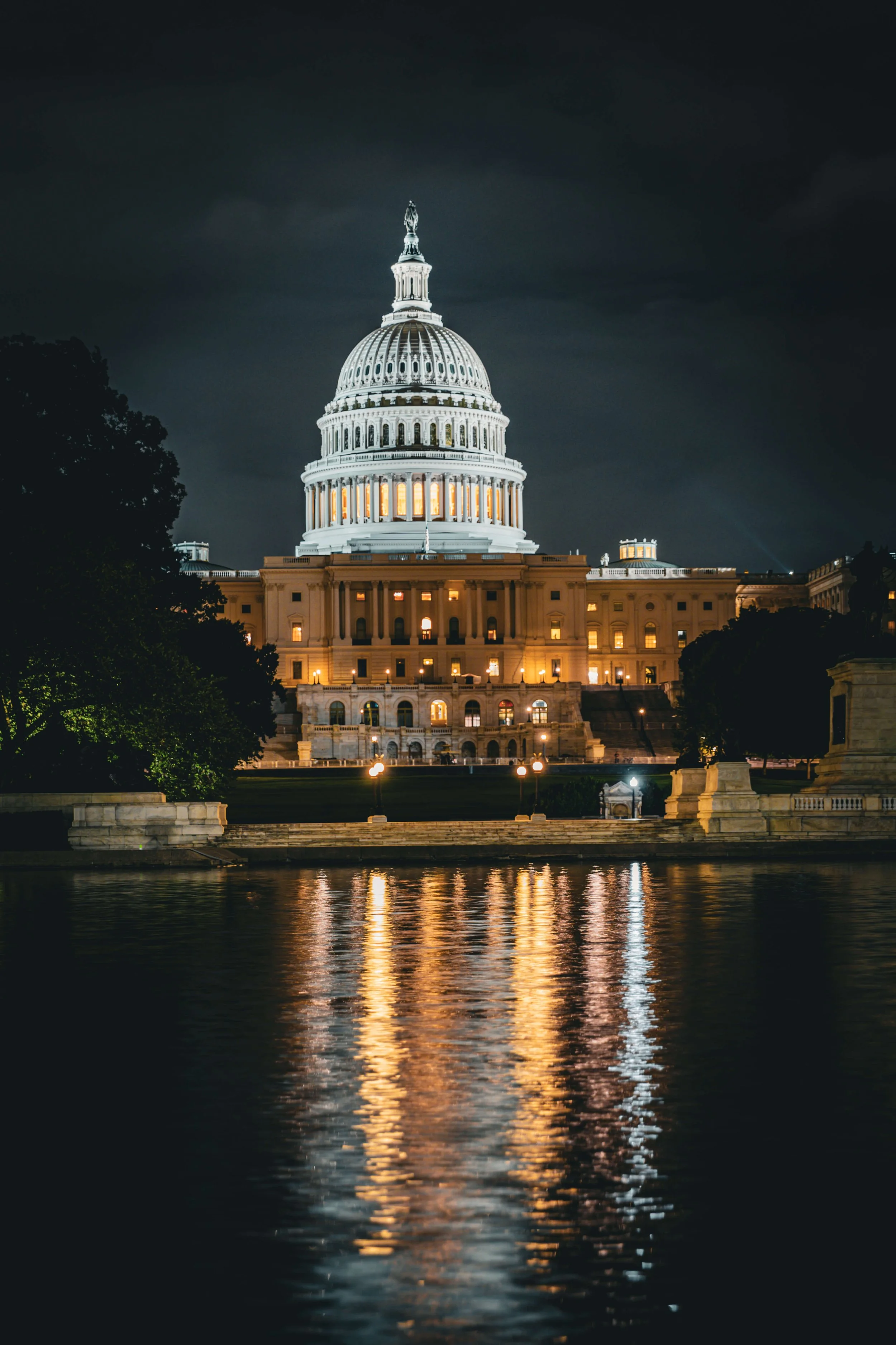 Nighttime view of the United States Capitol building in Washington, D.C., illuminated and reflecting in the water in the foreground.